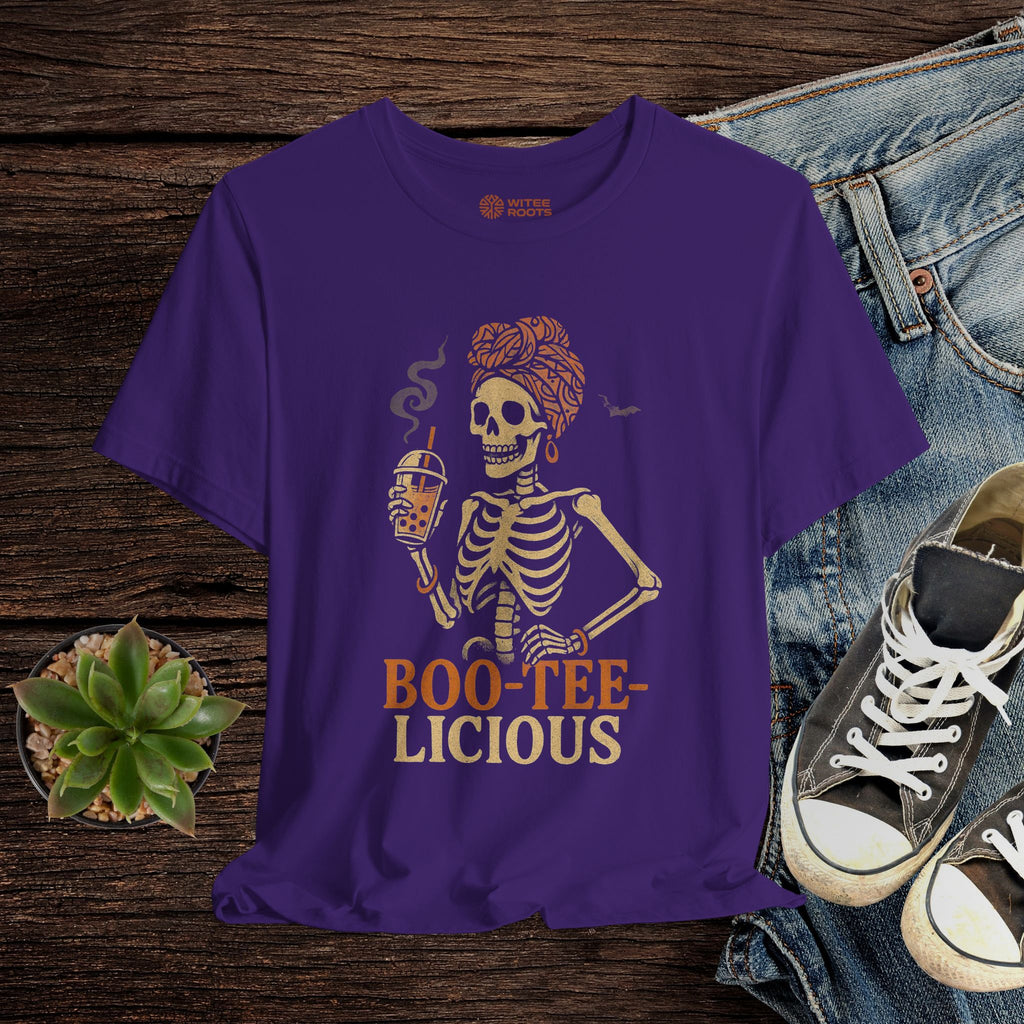 Purple t-shirt with skeleton design and 'BOO-TEE-LICIOUS' text on a wooden surface with jeans and sneakers.