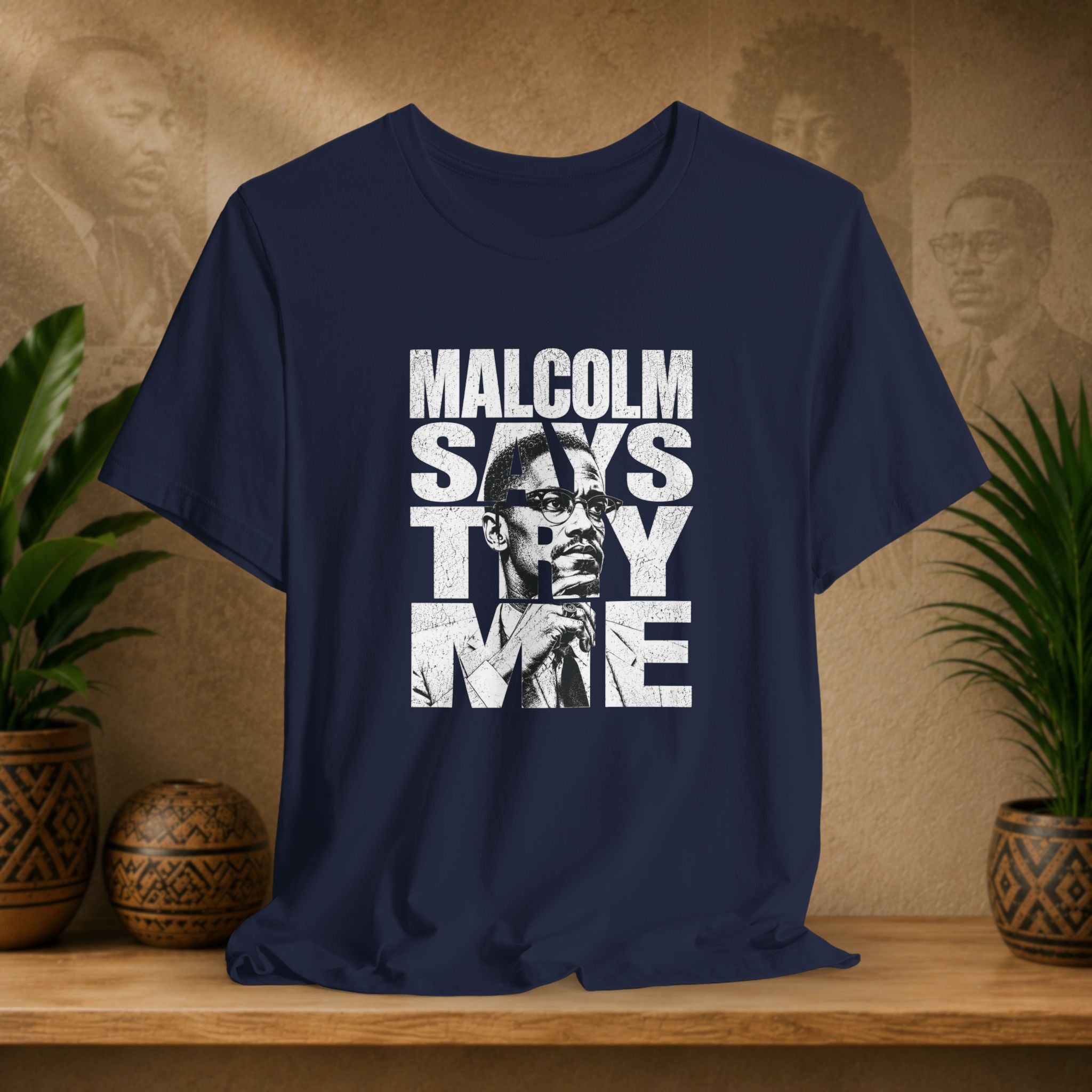 Navy blue t-shirt with a graphic design featuring a face and text on a wooden surface with plants and a textured wall in the background.