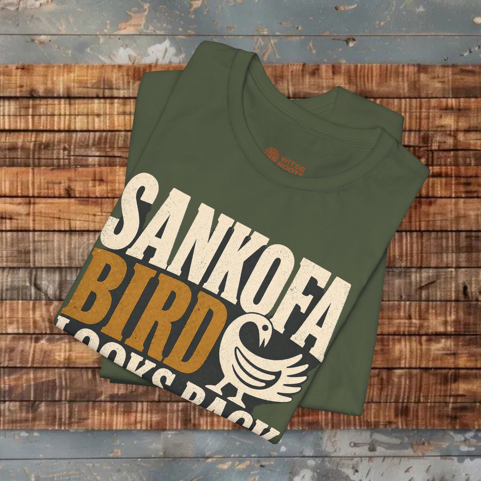 Folded green t-shirt with 'Sankofa Bird' text on a wooden surface