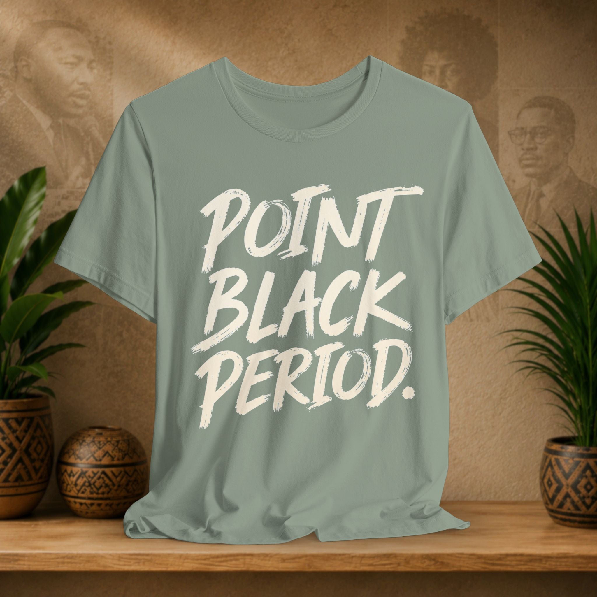 A sage green t-shirt with the text 'POINT BLACK PERIOD.' printed in a white brush-stroke font, displayed on a wooden shelf with potted plants and a textured background.