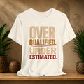 A beige short-sleeve graphic T-shirt displayed on a wooden surface, with the slogan 'OVER QUALIFIED. UNDER ESTIMATED.