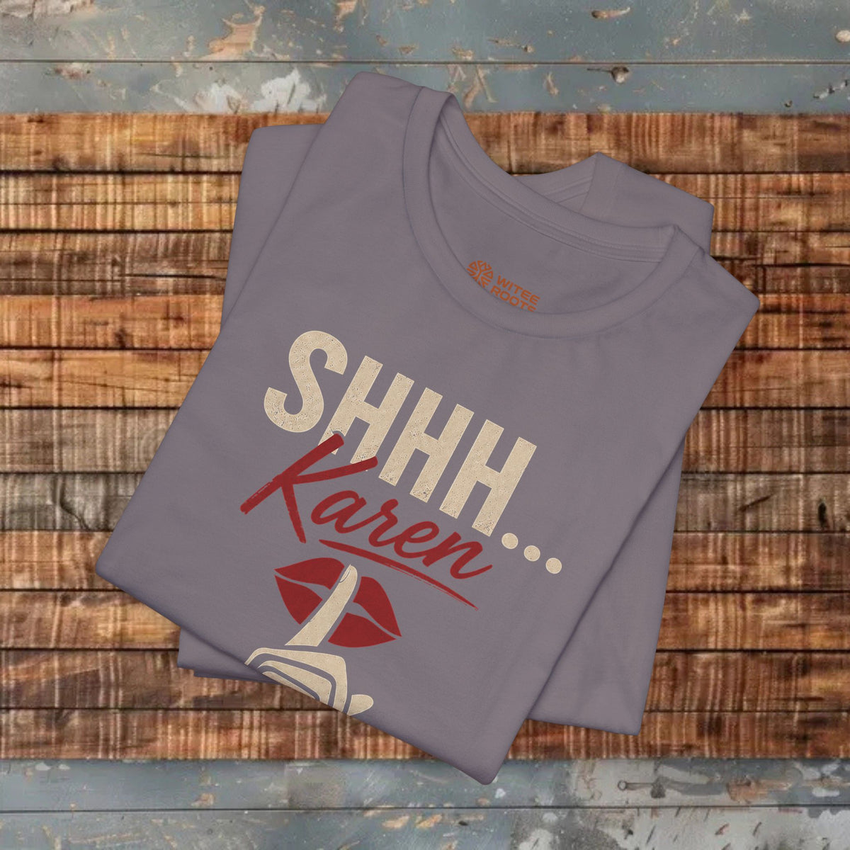 A folded gray t-shirt with a graphic print reading 'SHHH Karen' in beige and red, with red lips and a small white graphic below, displayed on a rustic wooden plank background.