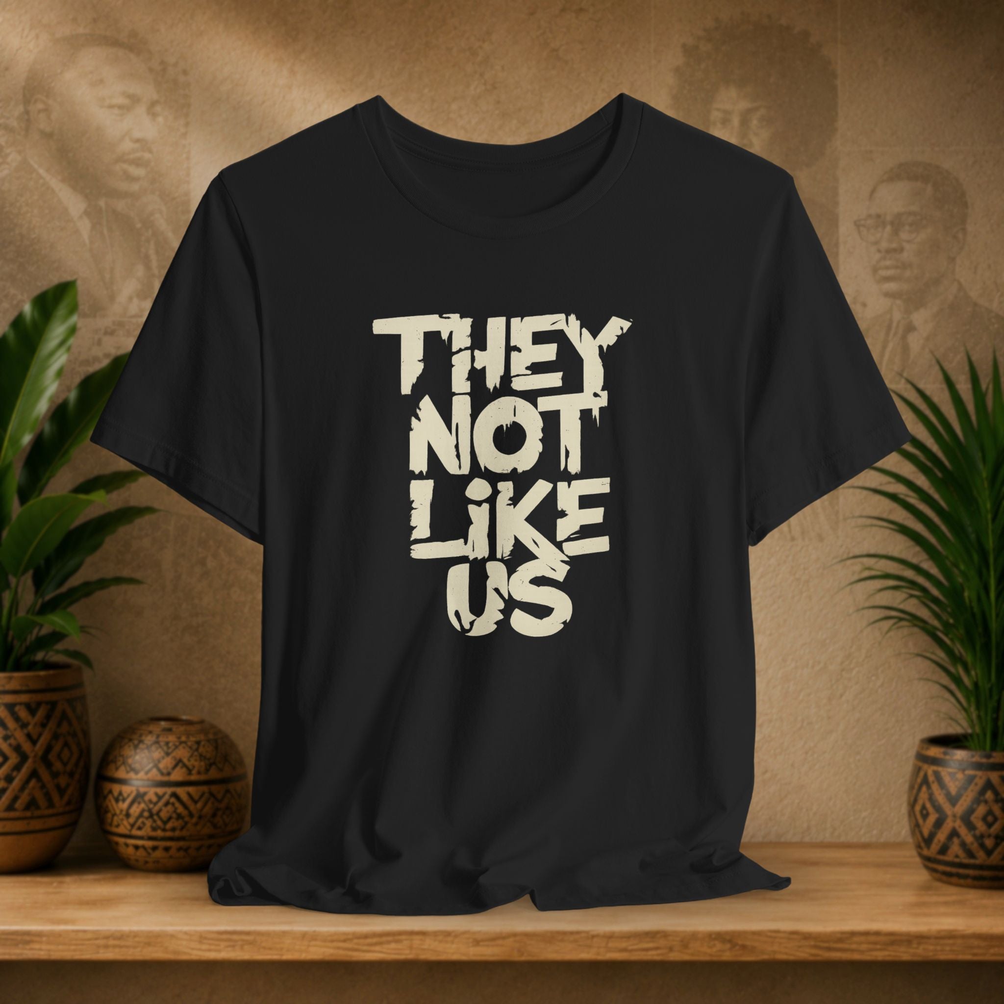 Black t-shirt with 'THEY NOT LIKE US' text on a wooden surface with plants and a textured wall background