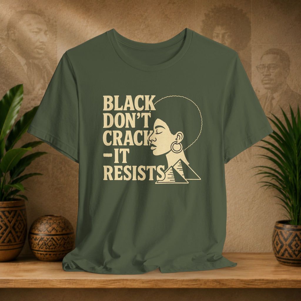 An olive green graphic t-shirt displayed on a wooden shelf, flanked by potted plants. The shirt features a cream-colored graphic with the text 'BLACK DON'T CRACK -IT RESISTS' alongside a profile illustration of a woman with an afro and geometric shapes.