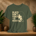 An olive green graphic t-shirt displayed on a wooden shelf, flanked by potted plants. The shirt features a cream-colored graphic with the text 'BLACK DON'T CRACK -IT RESISTS' alongside a profile illustration of a woman with an afro and geometric shapes.
