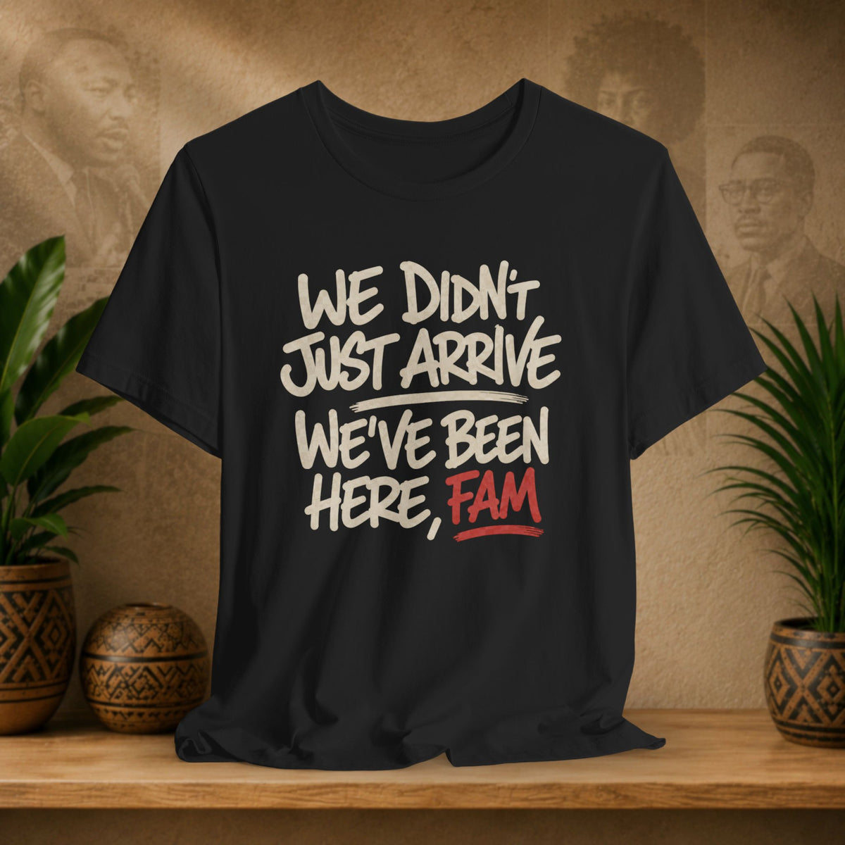 A black short-sleeve graphic T-shirt displayed on a wooden shelf, with white and red text reading “WE DIDN'T JUST ARRIVE WE'VE BEEN HERE, FAM.” The background is a warm beige wall with faint portraits and potted plants on both sides.