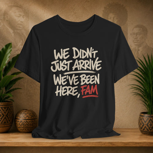 A black short-sleeve graphic T-shirt displayed on a wooden shelf, with white and red text reading “WE DIDN'T JUST ARRIVE WE'VE BEEN HERE, FAM.” The background is a warm beige wall with faint portraits and potted plants on both sides.