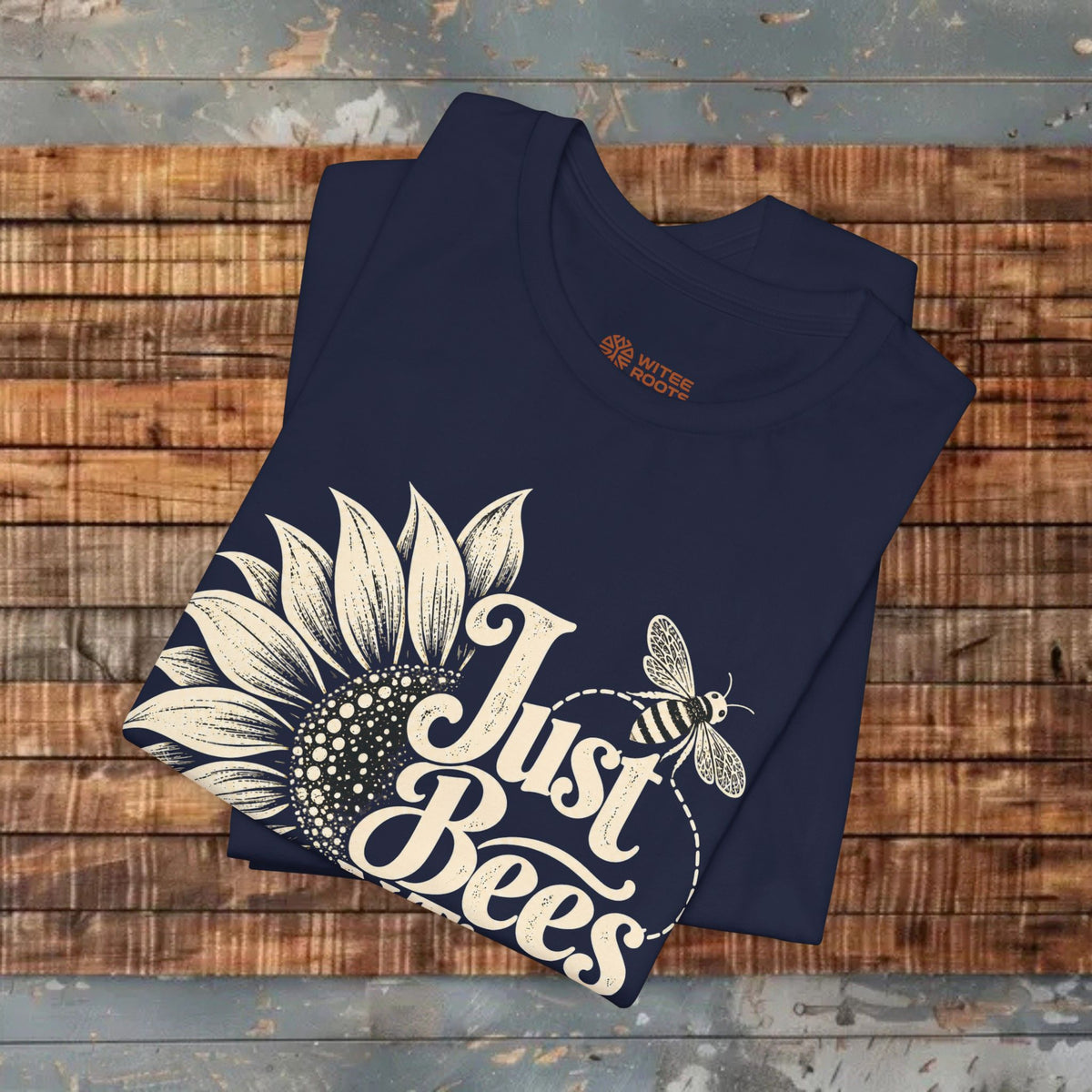 A navy blue folded t-shirt with a cream-colored sunflower and bee graphic that reads 'Just Bees' in script, displayed on a rustic wooden plank background.