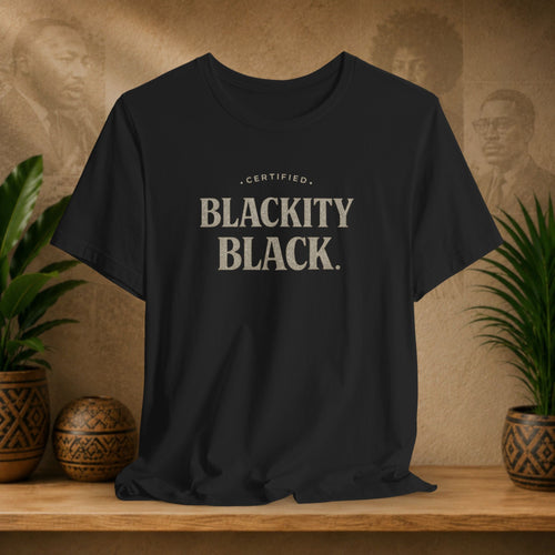 Black t-shirt with 'CERTIFIED BLACKITY BLACK' text on a wooden surface with plants and a vintage-style wall in the background.