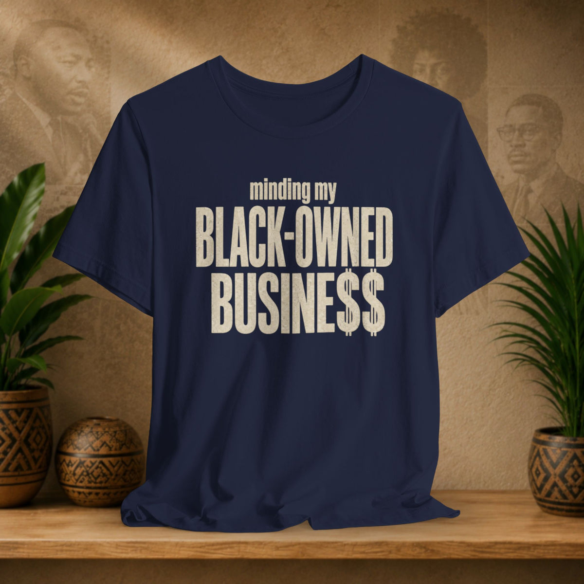 A navy t-shirt with a distressed cream-colored text graphic reading 'minding my BLACK-OWNED BUSINESS$$' displayed on a wooden shelf with decorative plants and textured background.