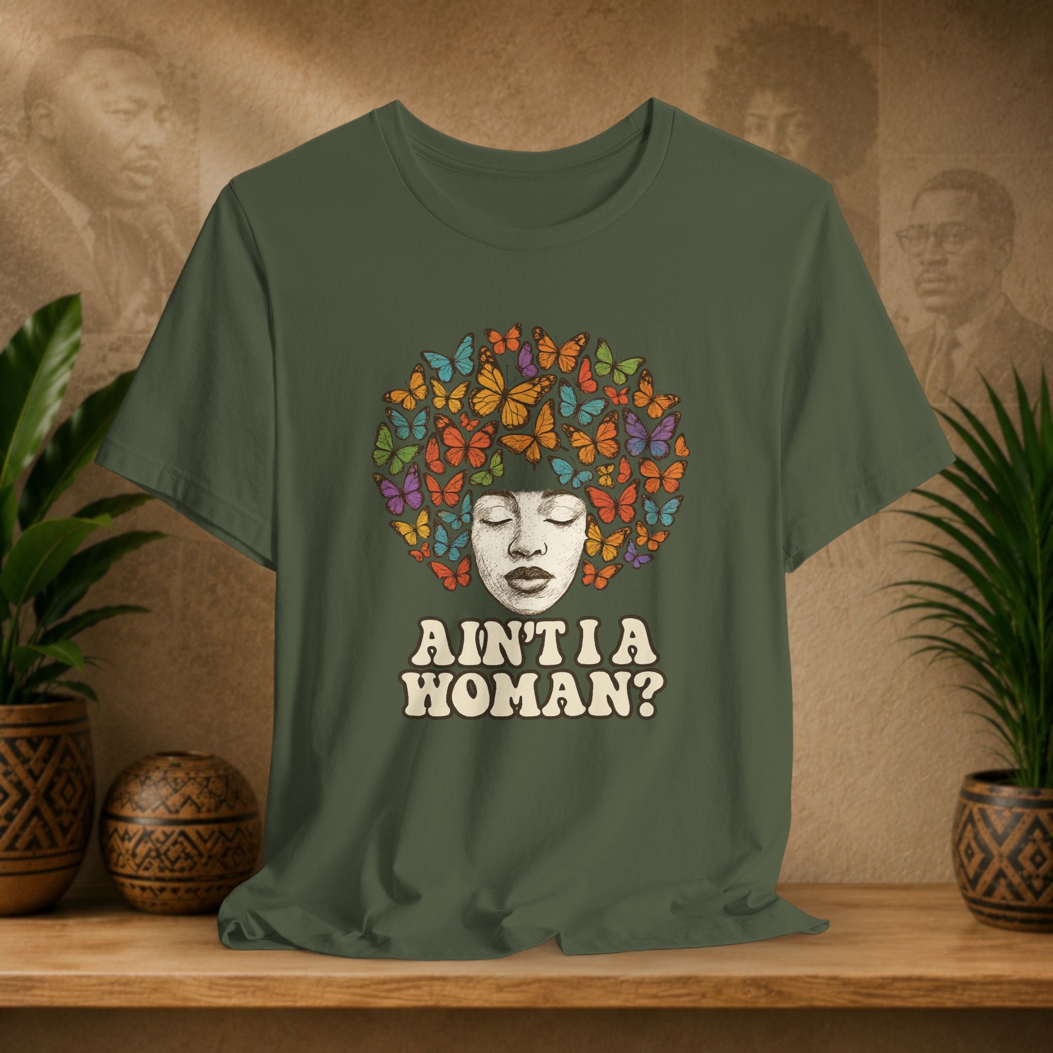 Green t-shirt with a colorful butterfly design and text on a wooden surface with plants and a textured wall in the background.