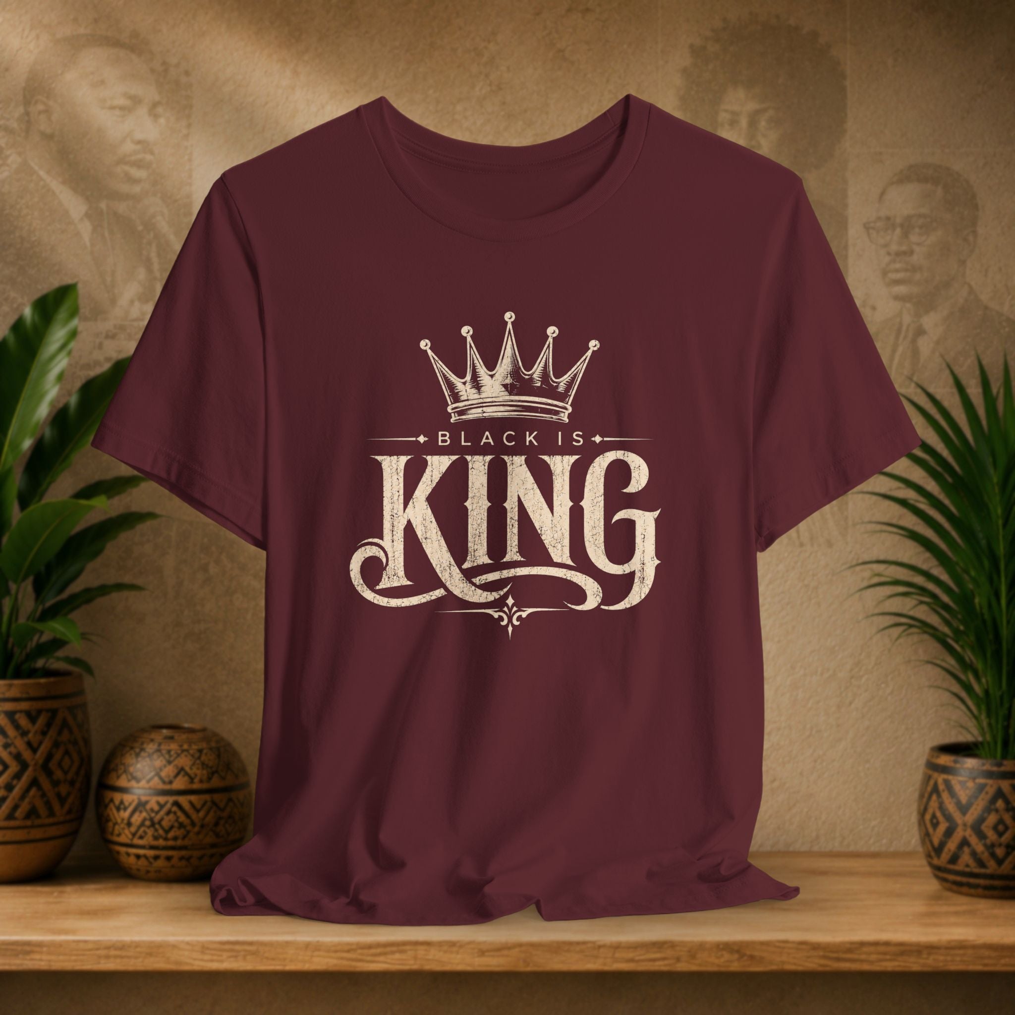 Maroon t-shirt with 'Black is King' design on a wooden surface with plants and decorative items in the background.