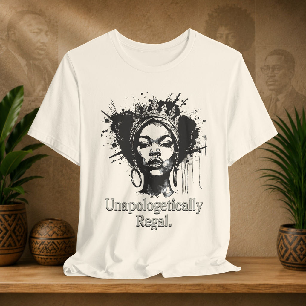 A beige short-sleeve graphic T-shirt displayed on a wooden surface, featuring a black illustrated portrait with paint splatter and the text 'Unapologetically Regal.' The shirt is photographed in a styled indoor setting with potted plants and a warm textured wall behind it.