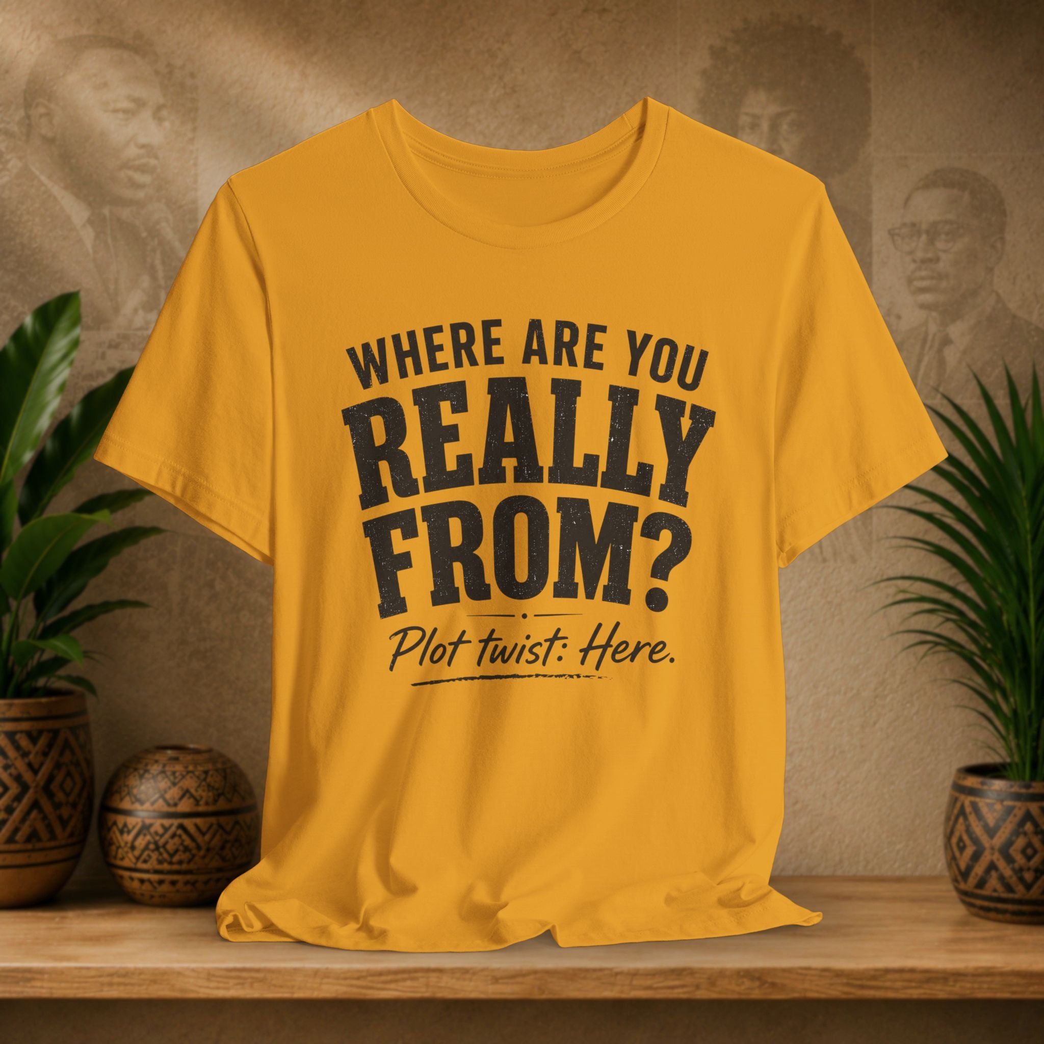 Yellow t-shirt with text 'WHERE ARE YOU REALLY FROM? Plot twist: Here.' on a wooden surface with decorative plants and background.