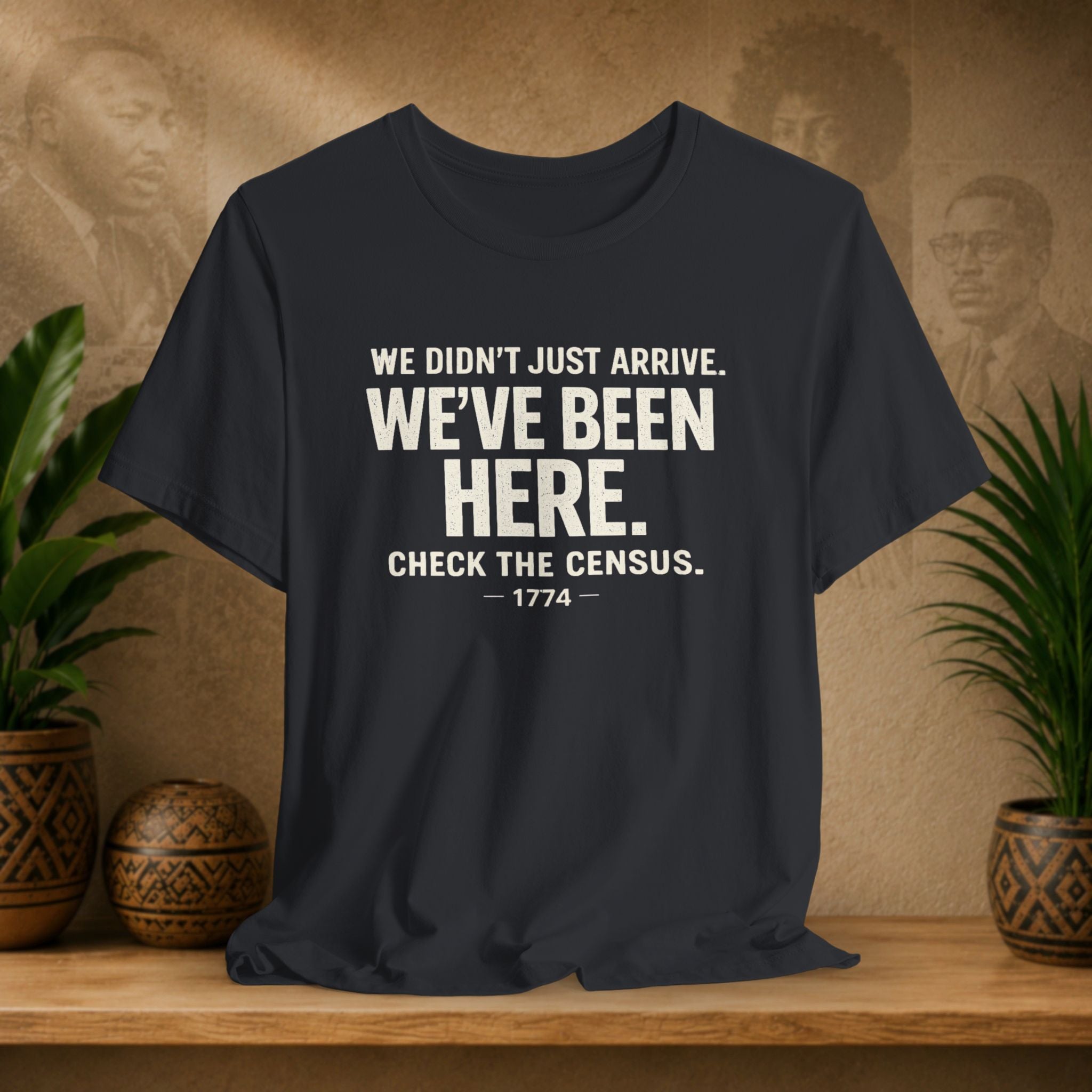 Black t-shirt with white text on a wooden surface with plants and a textured wall background