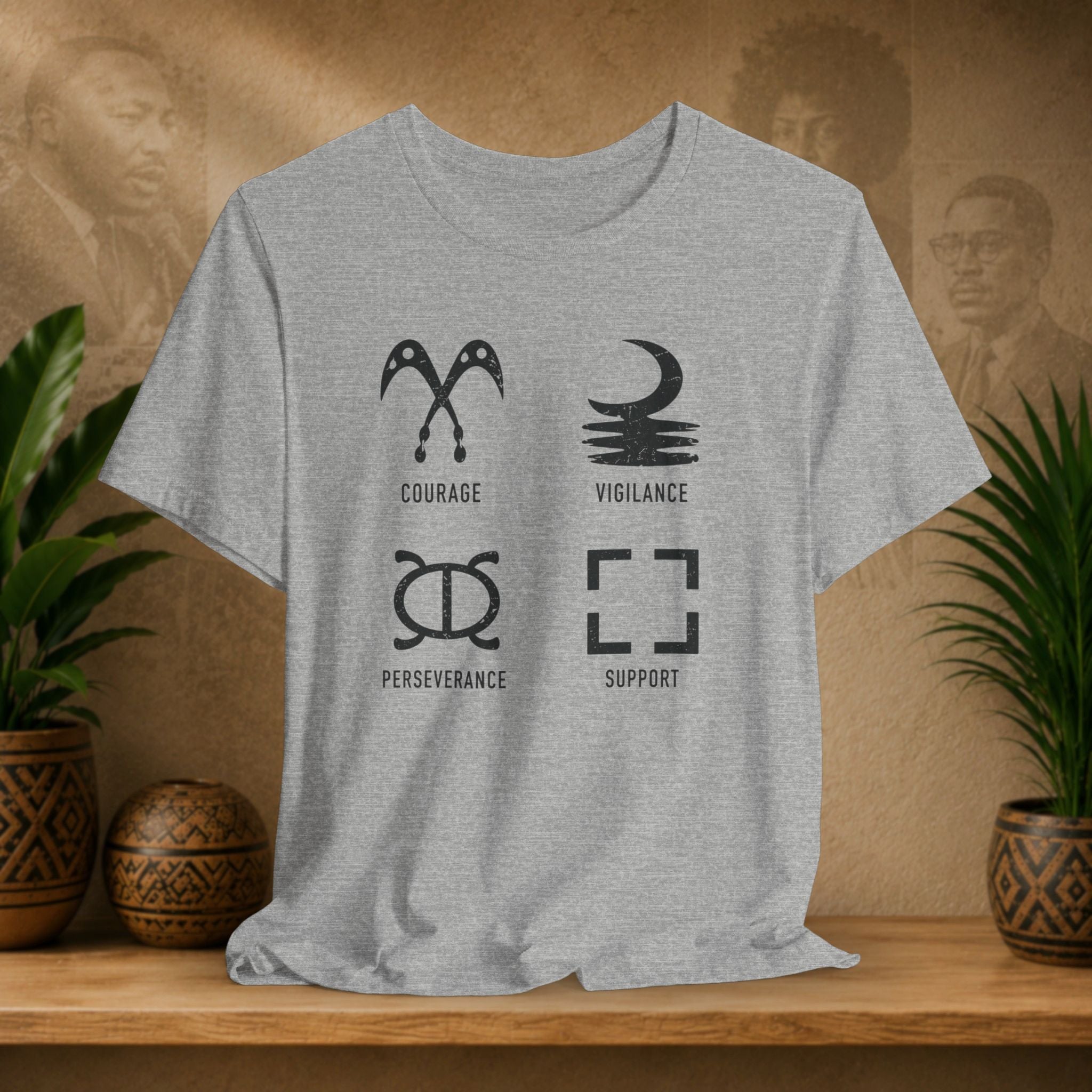 Gray t-shirt with black symbols and text on a wooden surface with plants and a textured wall background