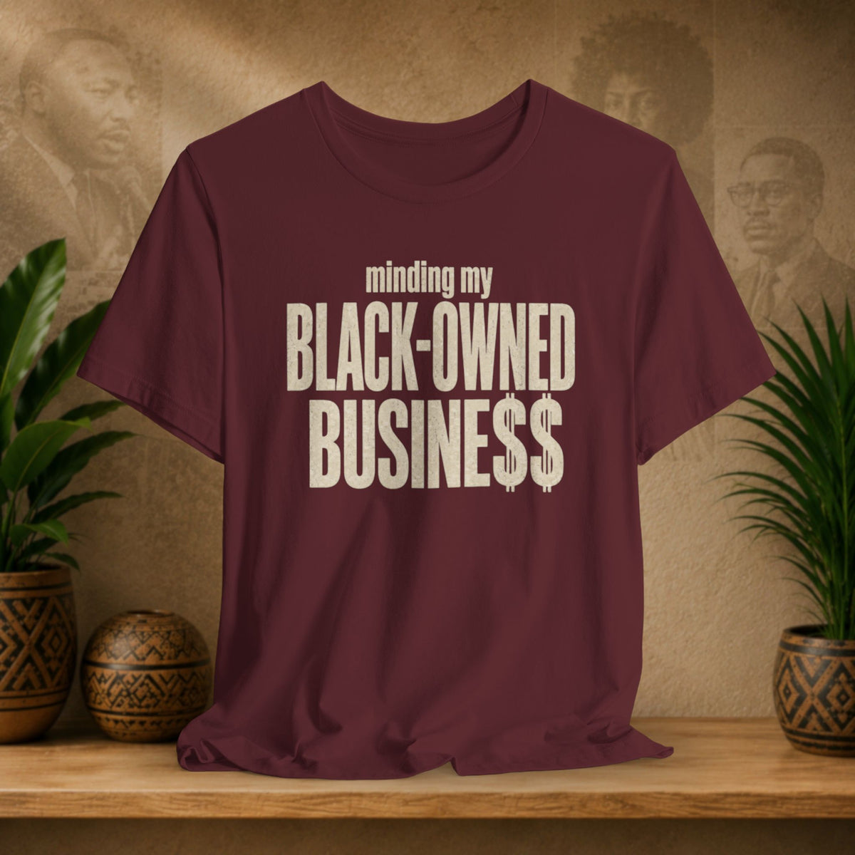 A maroon short-sleeve t-shirt with the text 'minding my BLACK-OWNED BUSINESS$$' printed in distressed off-white lettering on the front, displayed on a wooden shelf with decorative plants and textured background panels.