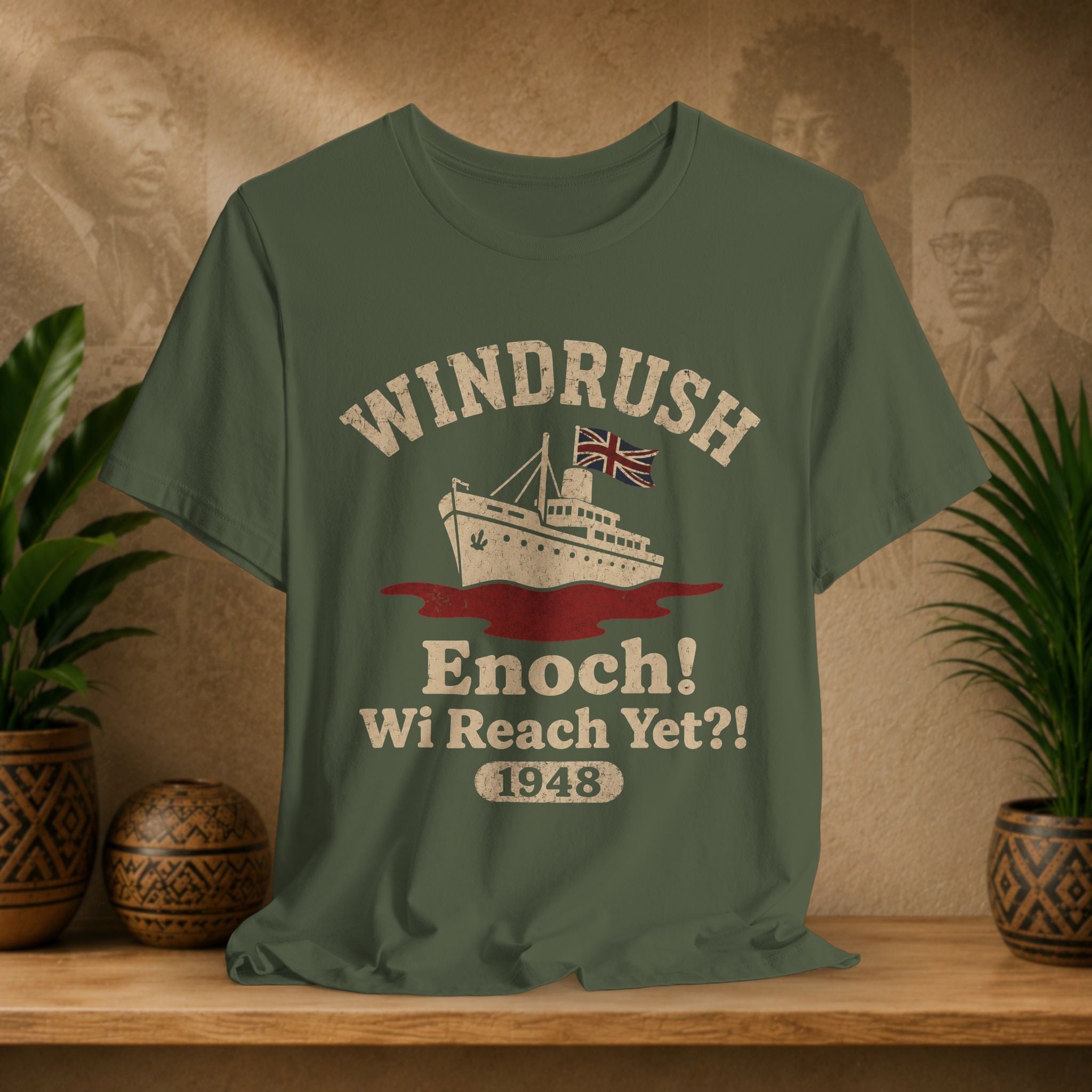 Green t-shirt with 'Windrush Enoch! Wi Reach Yet?! 1948' design on a wooden surface with plants and a decorative wall.