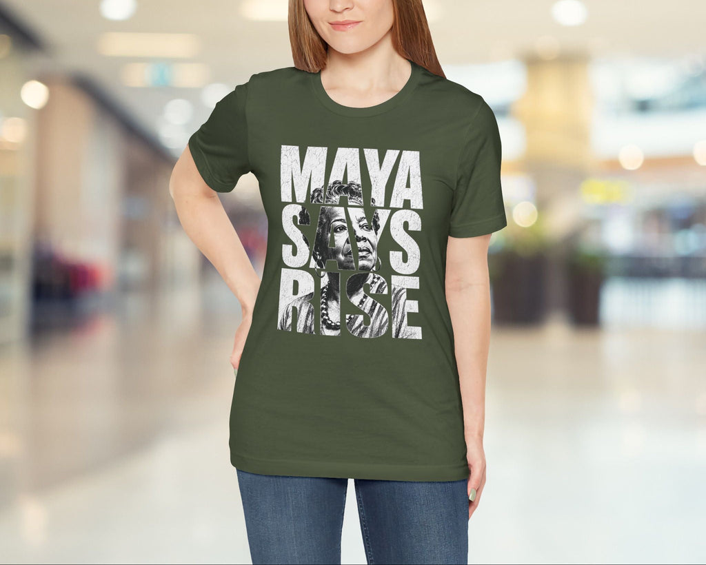 Woman wearing a green t-shirt with 'MAYA SAYS RISE' text and graphic in a blurred indoor setting