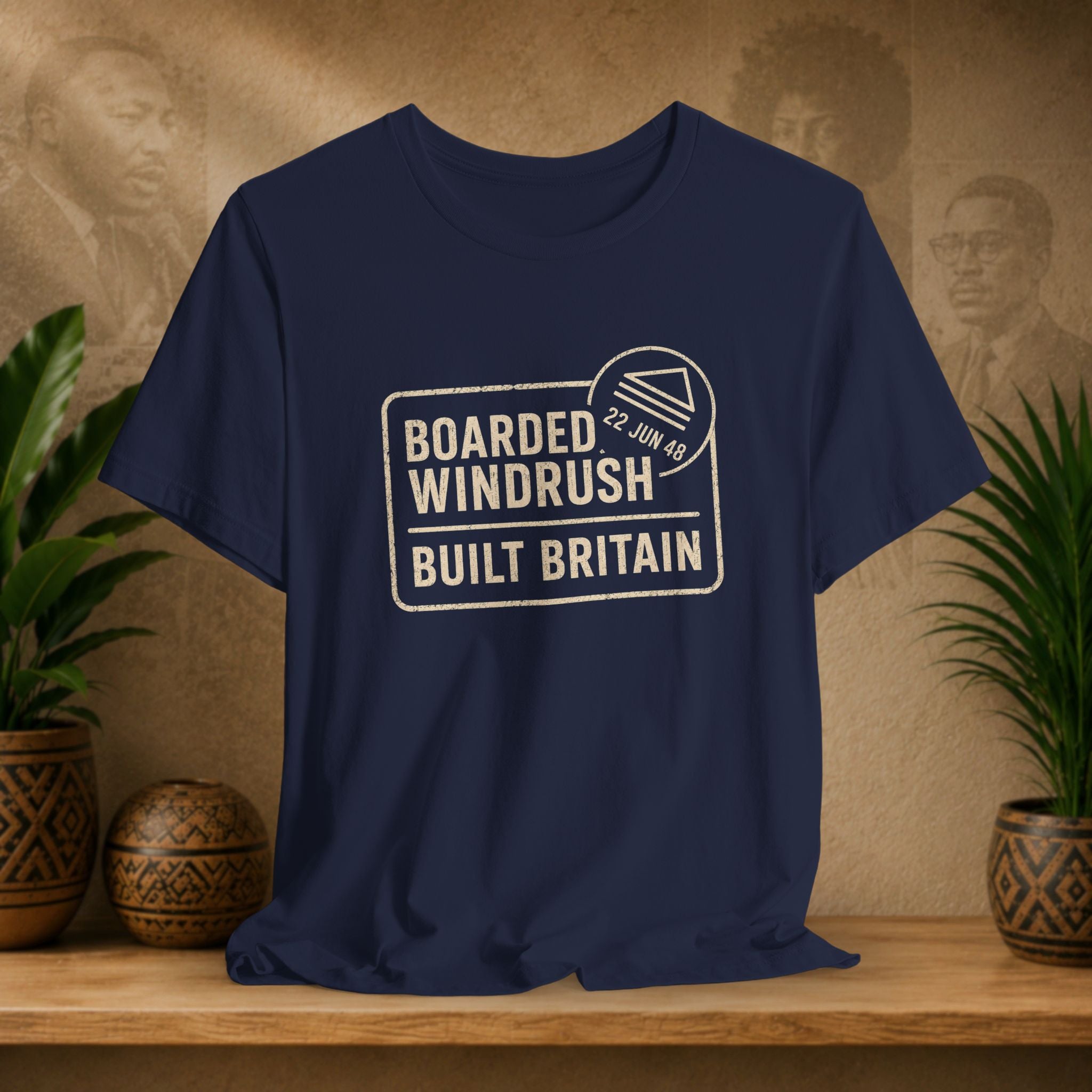 Navy blue t-shirt with 'Boarded Windrush Built Britain' text on a wooden surface with plants and a textured wall background.