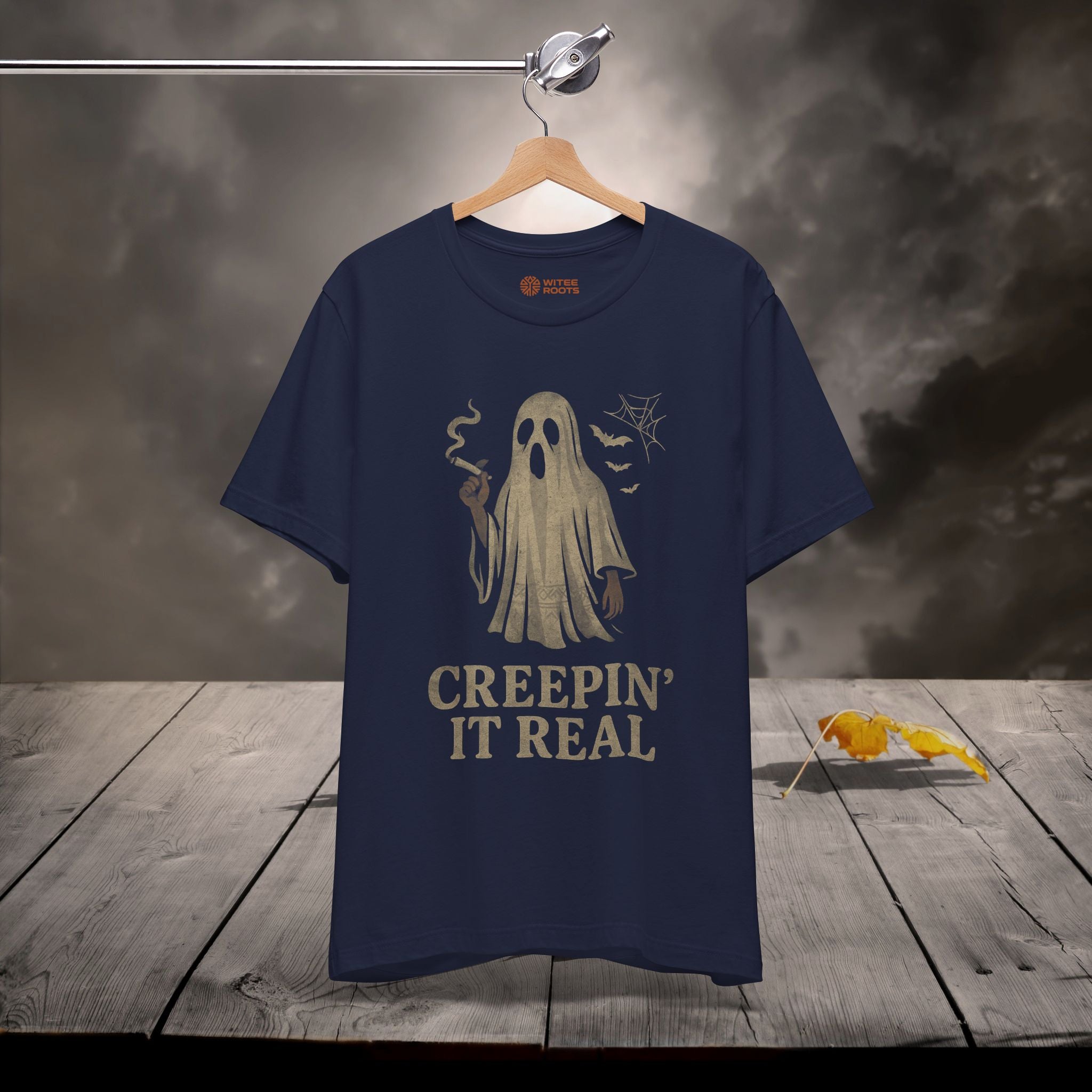 A navy blue graphic t-shirt hanging on a wooden hanger against a dark, moody background with a rustic wooden table. The shirt features a graphic of a ghost holding a candle with the text 'CREEPIN' IT REAL' printed below in a distressed beige font.
