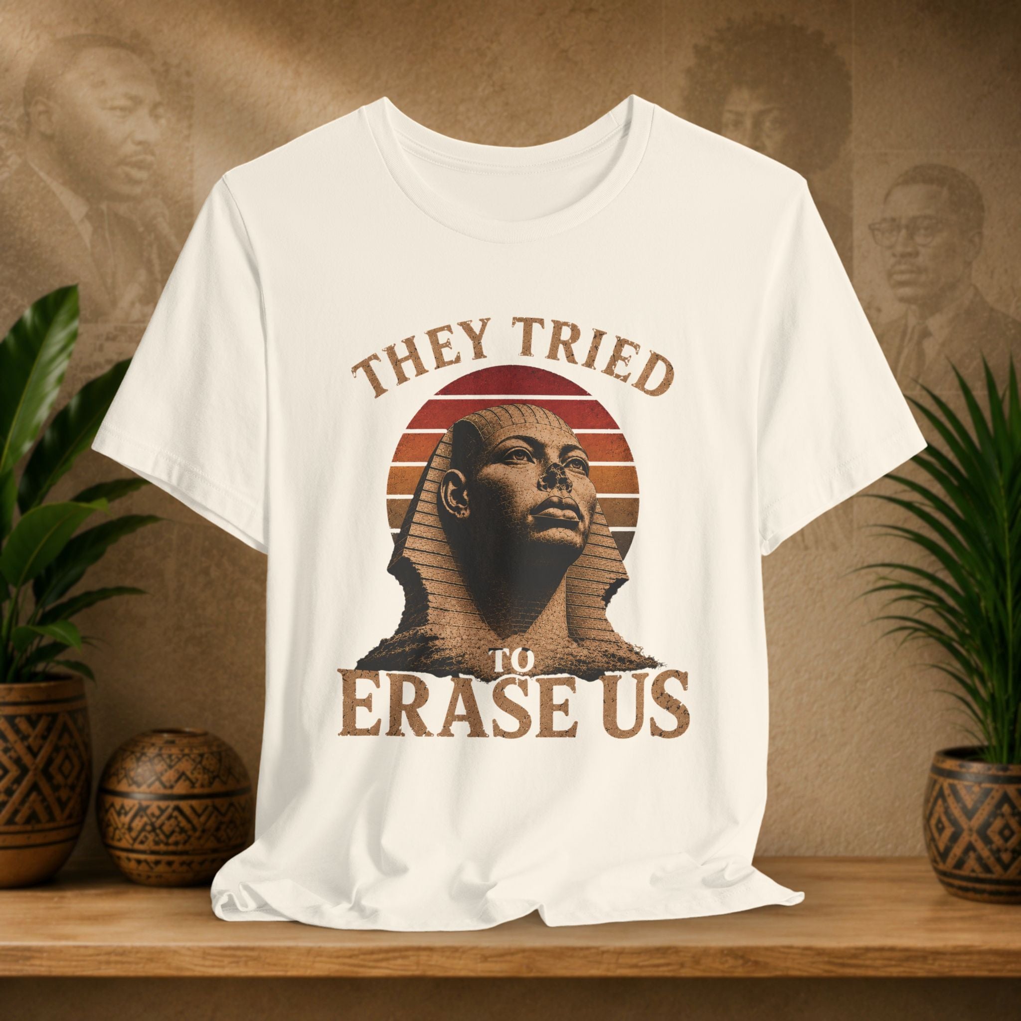 White t-shirt with a graphic of a sphinx and text 'They Tried To Erase Us' on a wooden surface with plants and a textured wall background.