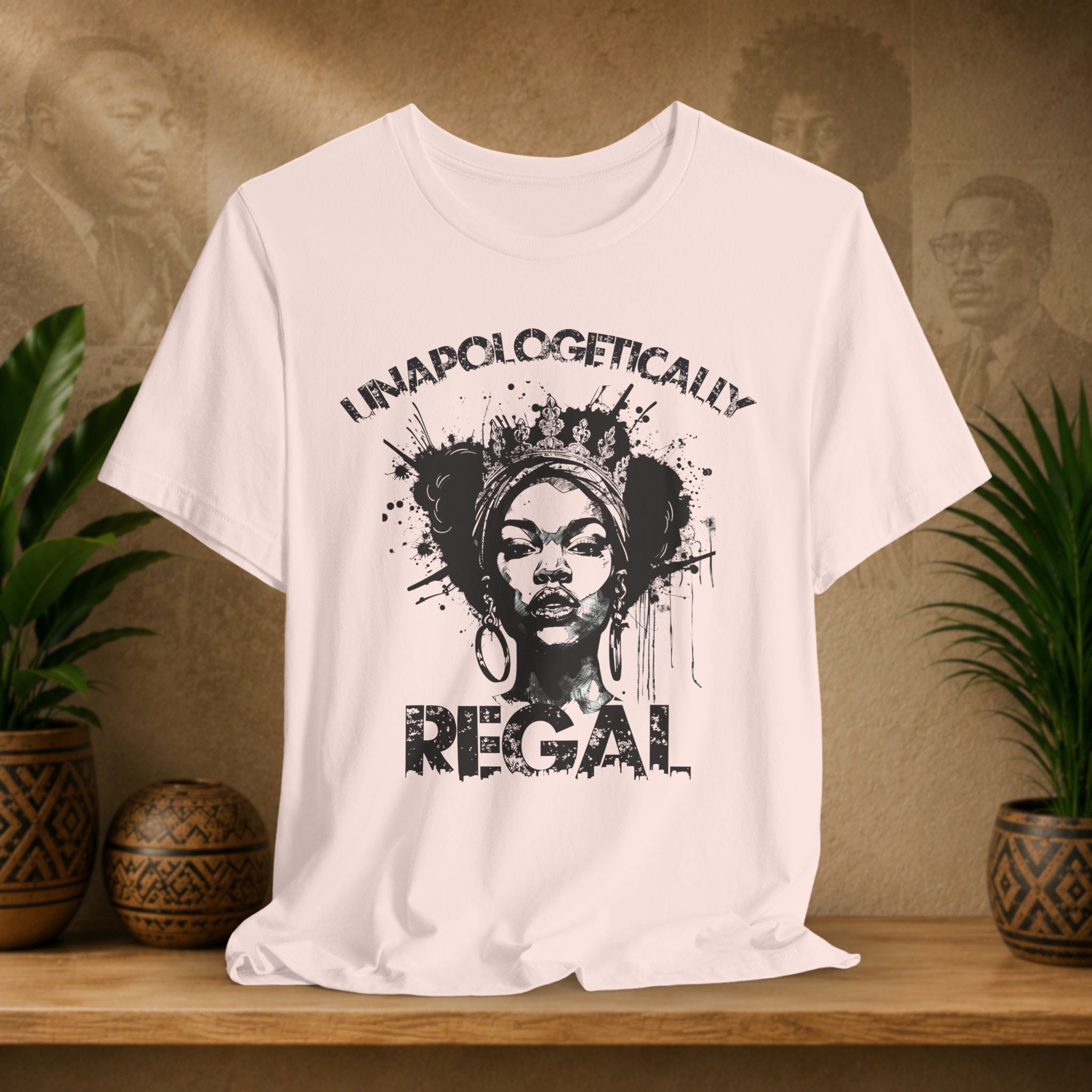 A soft pink short-sleeve graphic T-shirt displayed on a wooden surface, featuring a black distressed print of a woman's portrait with the words 'UNAPOLOGETICALLY REGAL.' The shirt is styled in a warm indoor setting with potted plants and a textured wall in the background.