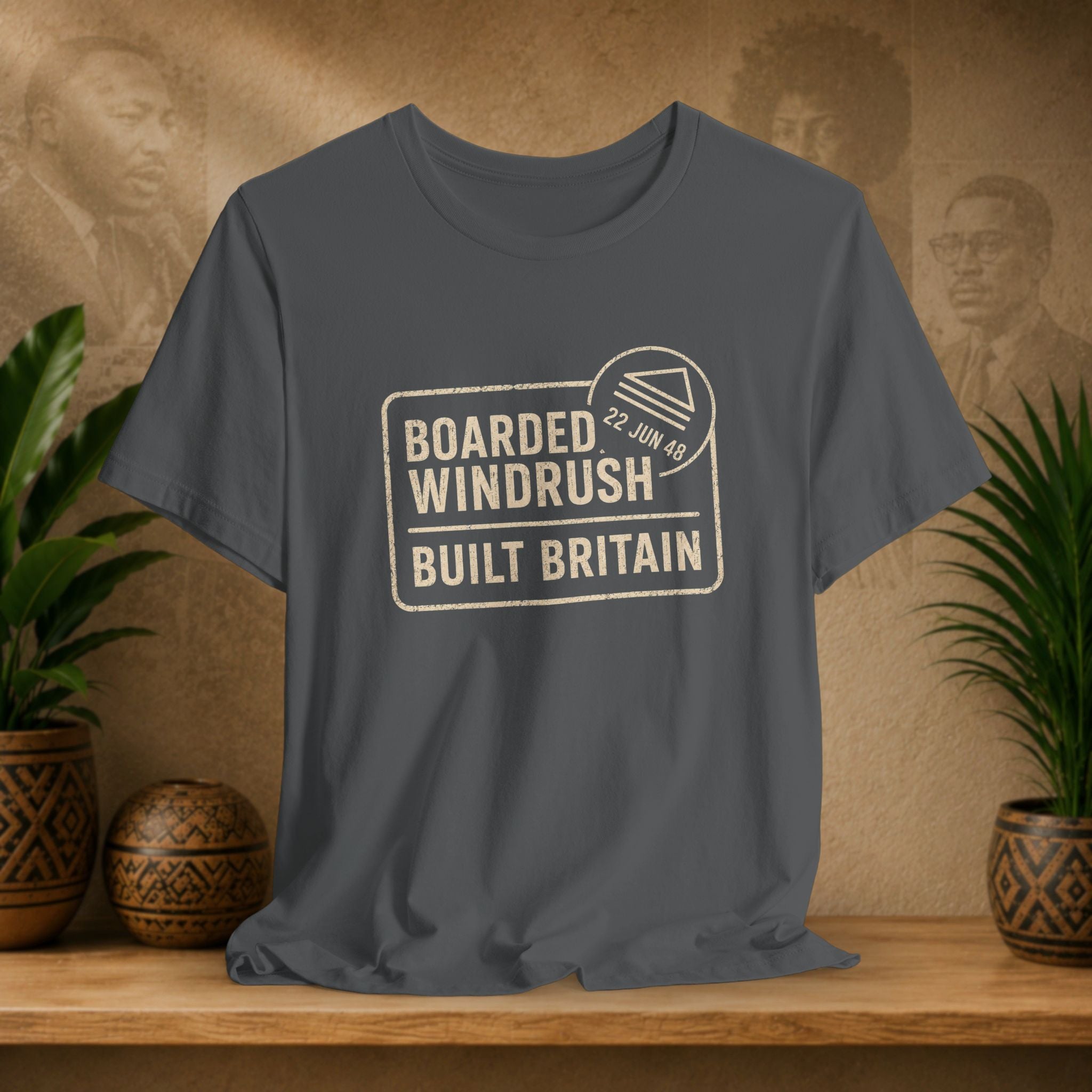 Gray t-shirt with 'Boarded Windrush Built Britain' text on a wooden surface with plants and a textured wall background