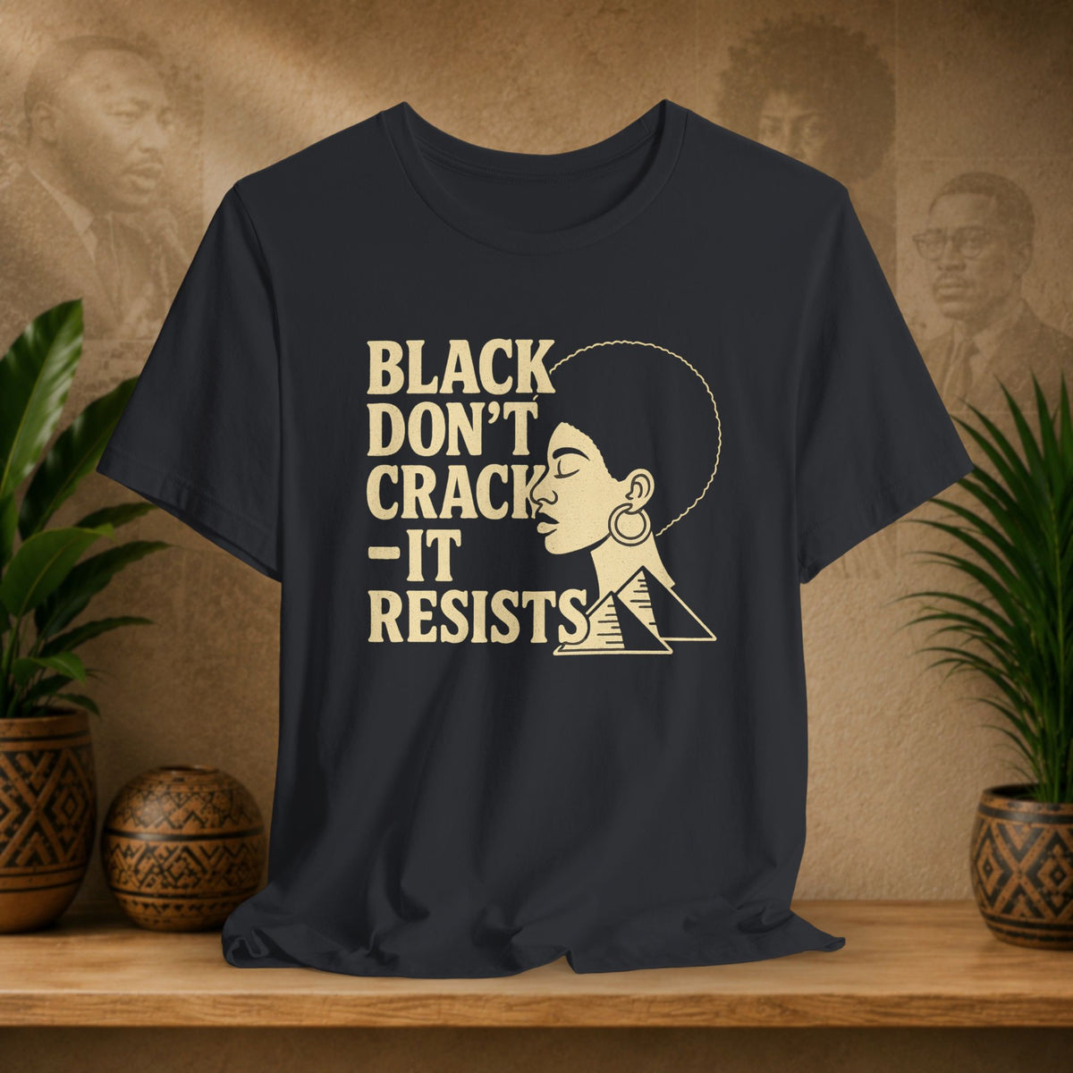 A black graphic t-shirt displayed on a wooden shelf, flanked by potted plants. The shirt features a beige graphic of a woman's profile next to the text 'BLACK DON'T CRACK - IT RESISTS'.