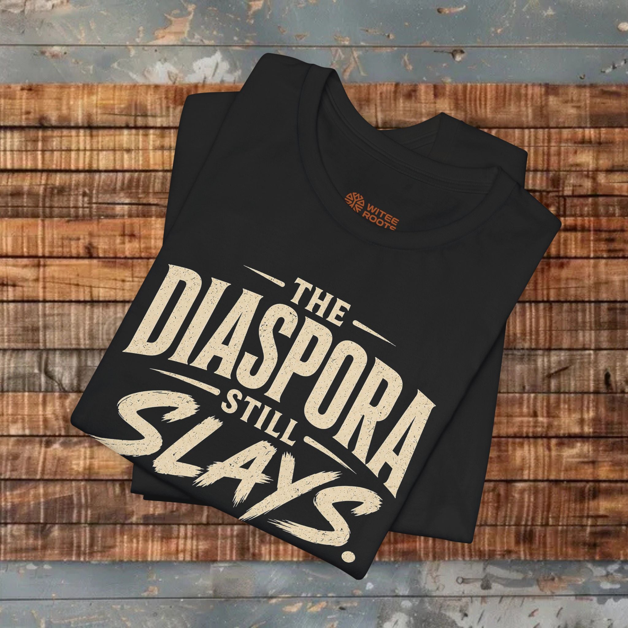 Black folded t-shirt with 'The Diaspora Still Slays Every Generation' text on a wooden background.