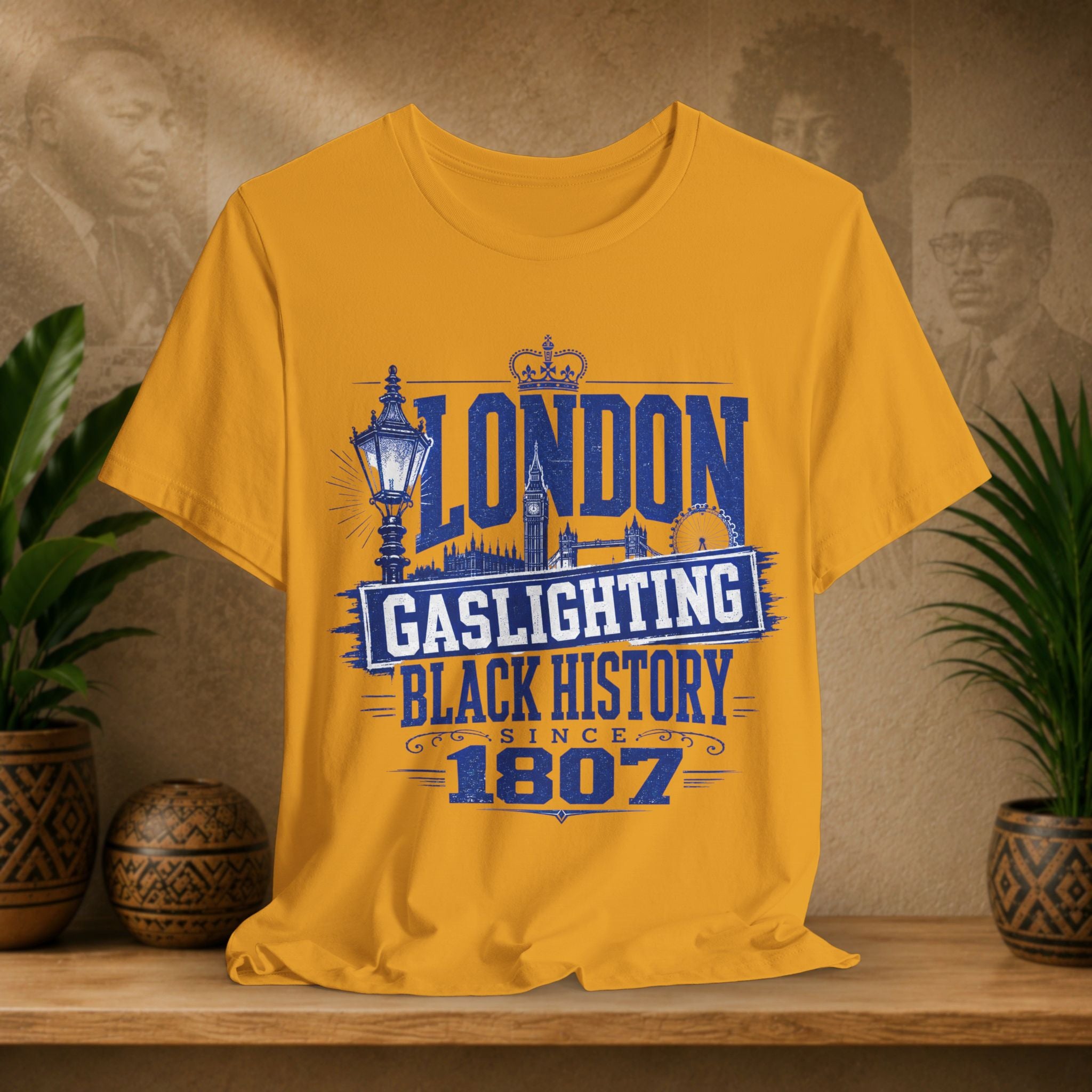 Mustard t-shirt with 'London Gaslighting Black History Since 1807' design on a wooden surface with plants and a decorative wall.