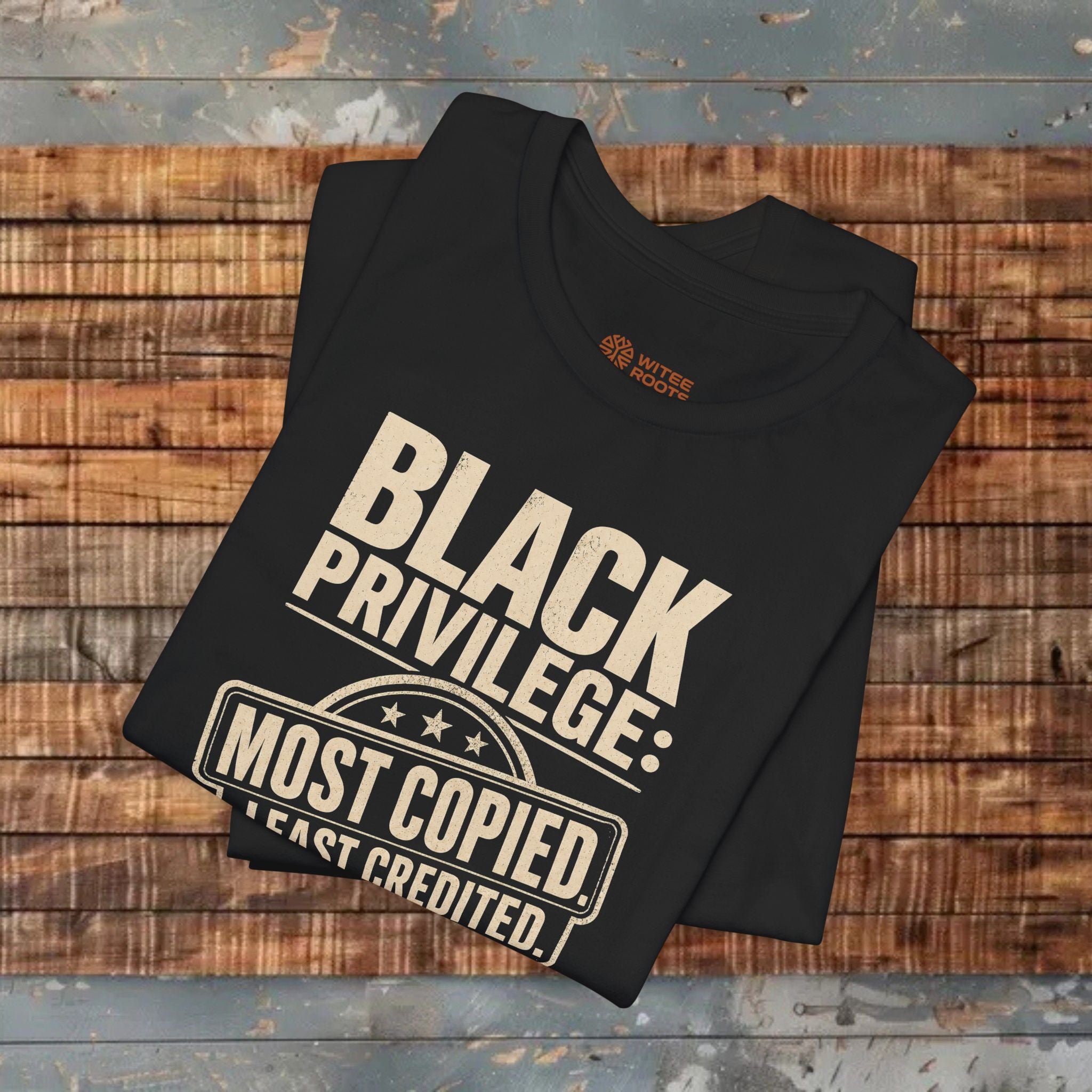 Black t-shirt with 'Black Privilege: Most Copied, Least Credited' text on a wooden surface