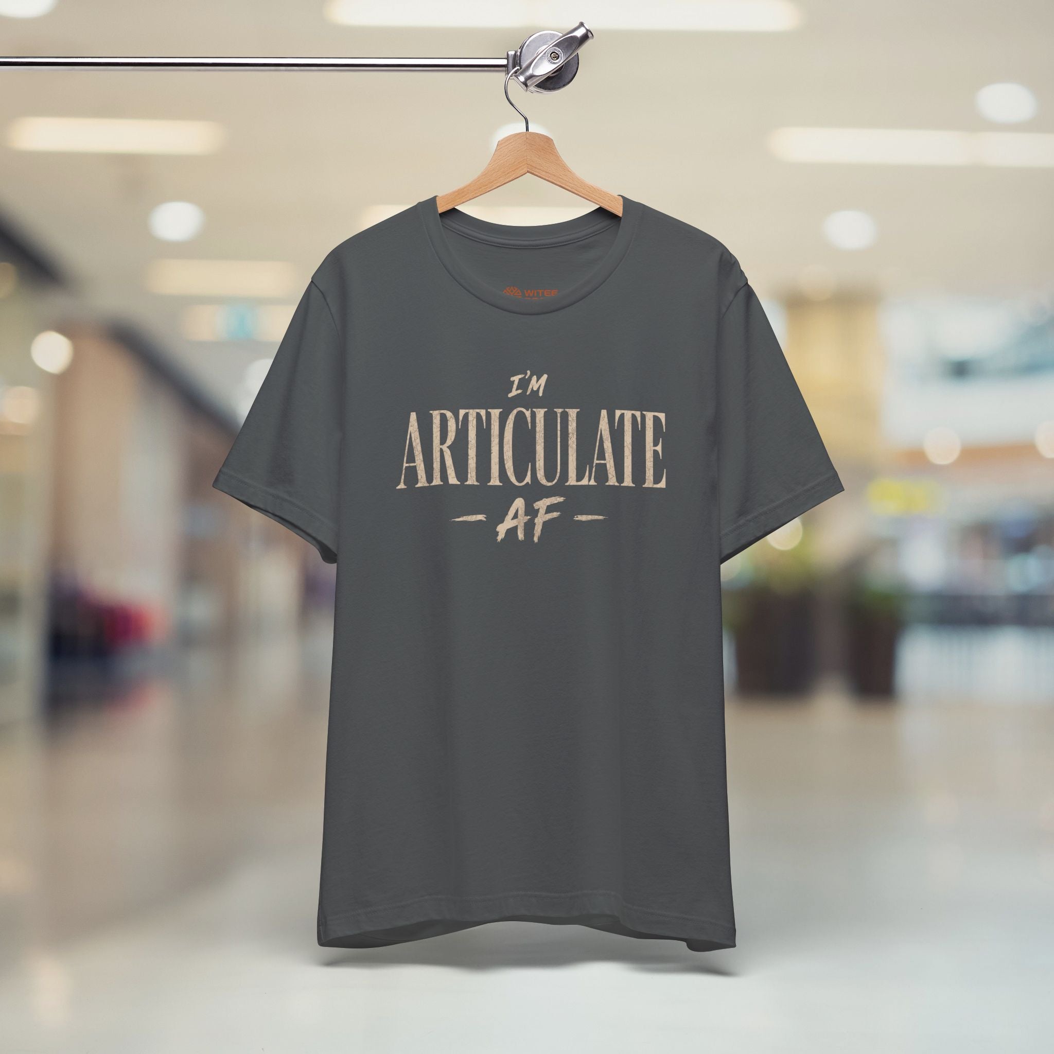 A dark gray crewneck t-shirt with the text 'I'M ARTICULATE AF' printed in a distressed beige font, displayed on a blurred shopping mall background.