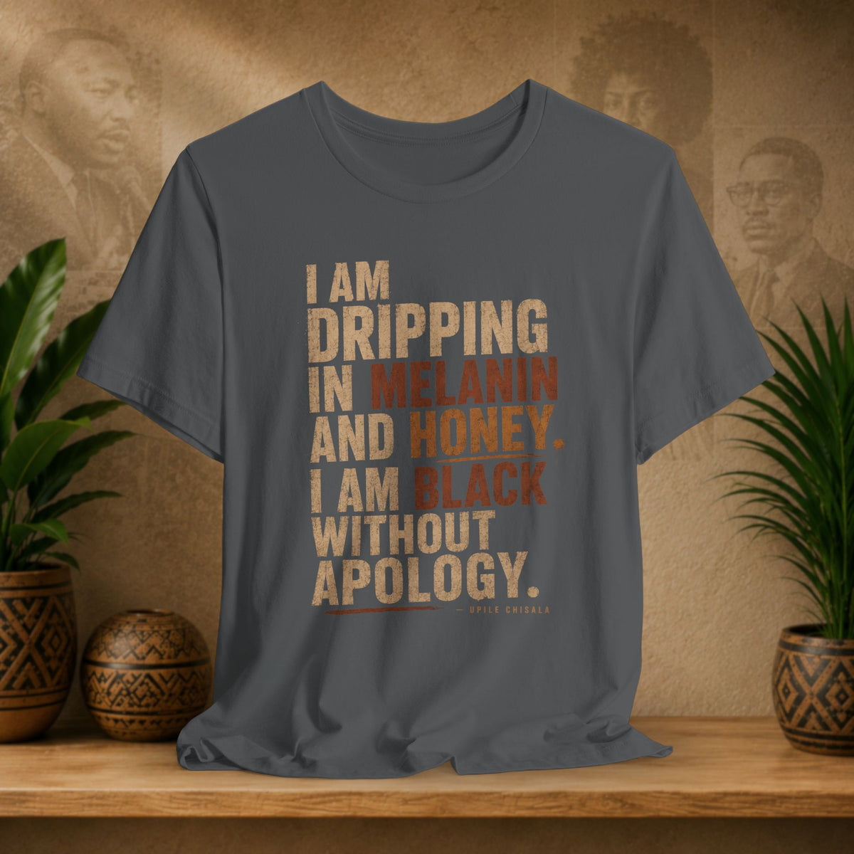 A dark gray graphic t-shirt featuring a quote by Little Rock Brown in distressed beige and orange lettering, displayed on a wooden shelf with potted plants and a textured background.
