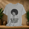 Gray t-shirt with a graphic of a woman with an afro and text 'No Justice No Peace No Quiet' on a wooden surface with plants and a textured wall in the background.
