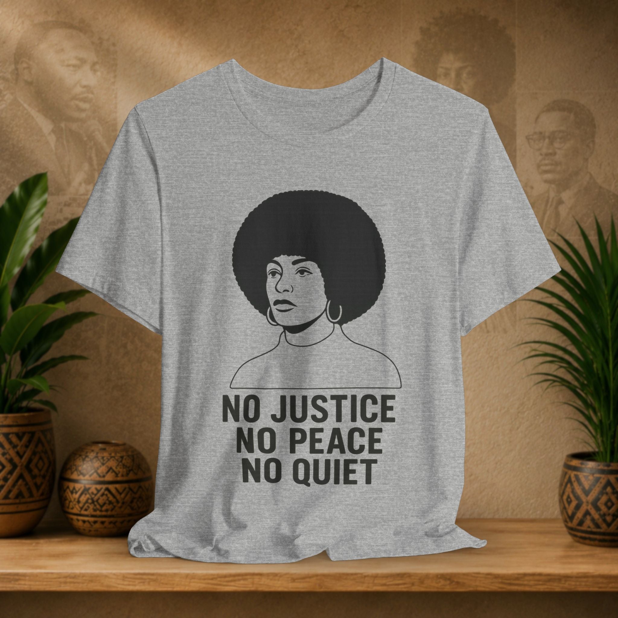 Gray t-shirt with a graphic of a woman with an afro and text 'No Justice No Peace No Quiet' on a wooden surface with plants and a textured wall in the background.