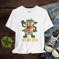 White t-shirt with a cartoon beer can design and 'Yes We Can' text, displayed with jeans and sneakers on a wooden surface.