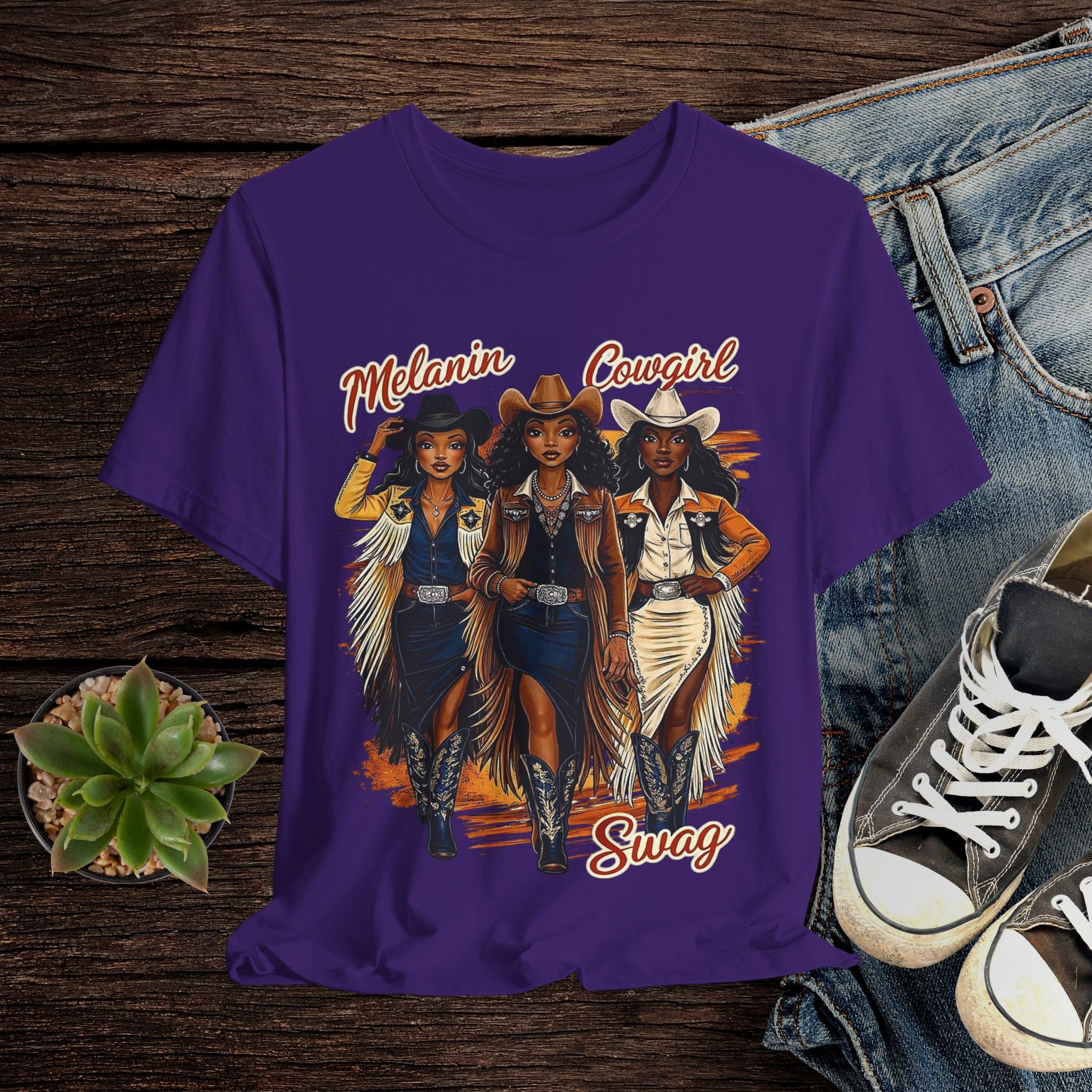 Purple t-shirt with cowgirl graphic and text on a wooden surface with jeans and sneakers.