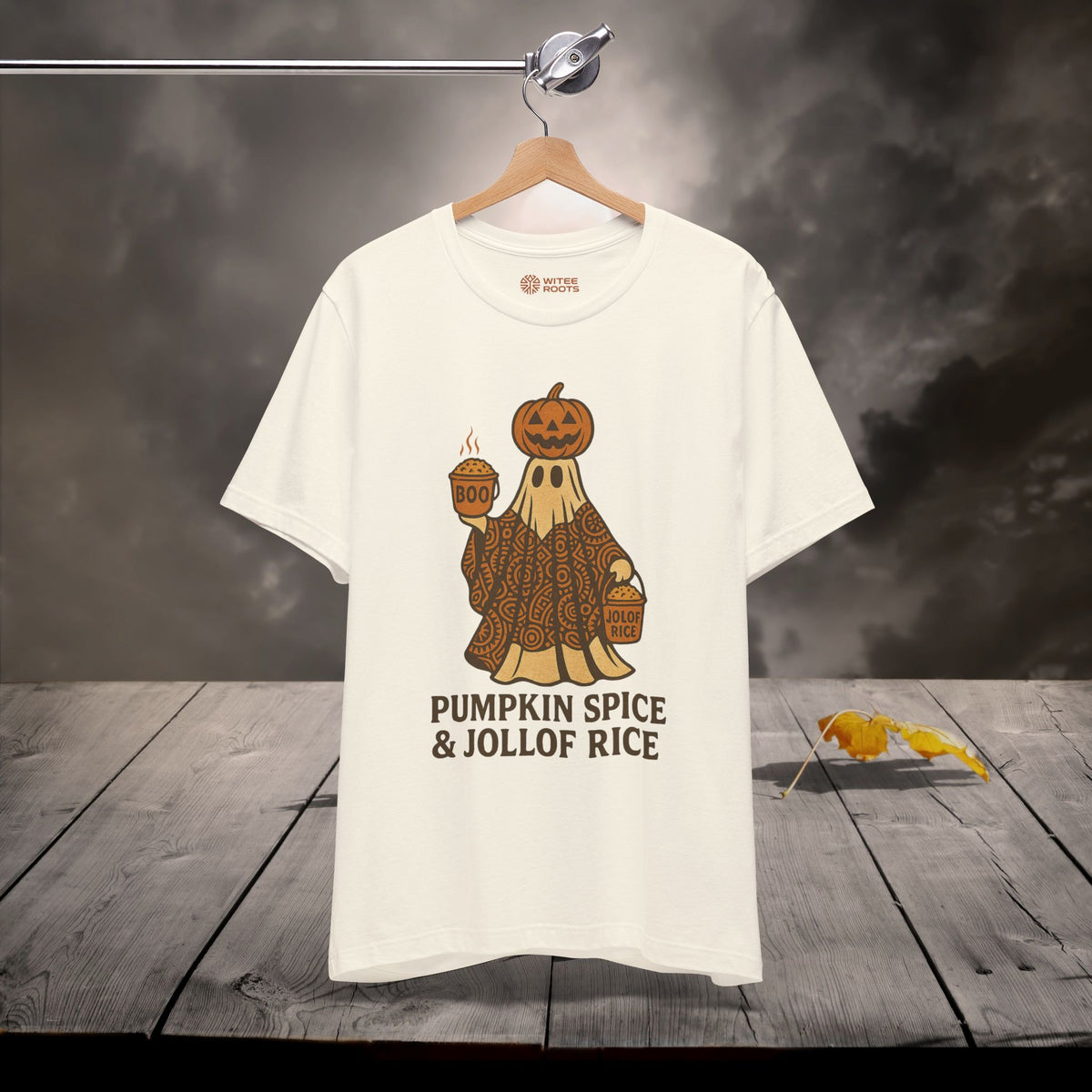 A white graphic t-shirt hanging on a wooden hanger against a dark, smoky background with a rustic wooden table below. The graphic features a cartoon pumpkin spice ghost wearing a patterned robe and holding a mug, with the text 'PUMPKIN SPICE & JOLLOF RICE' printed underneath.
