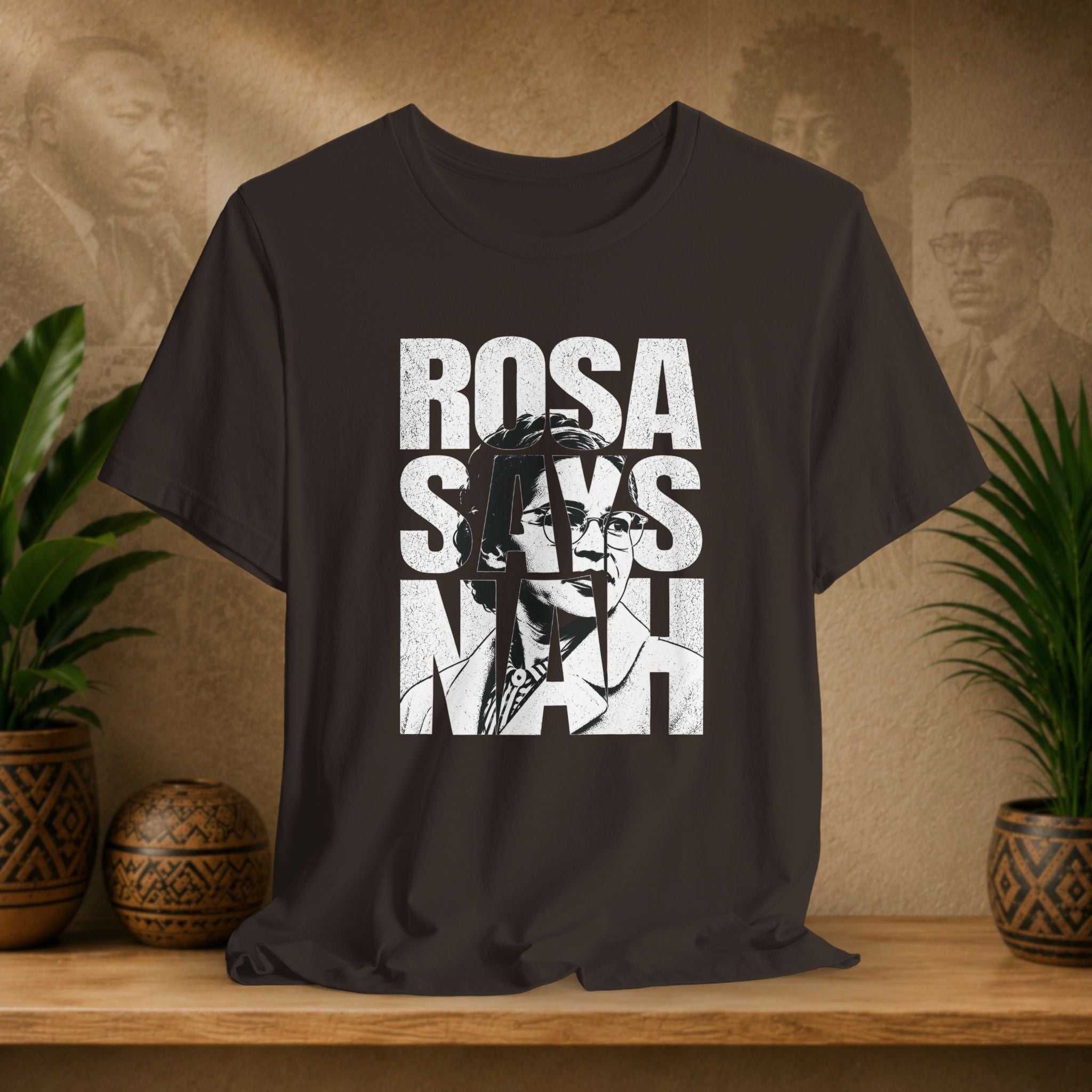 Black t-shirt with 'Rosa Parks' graphic design on a wooden surface with plants and a textured wall in the background.