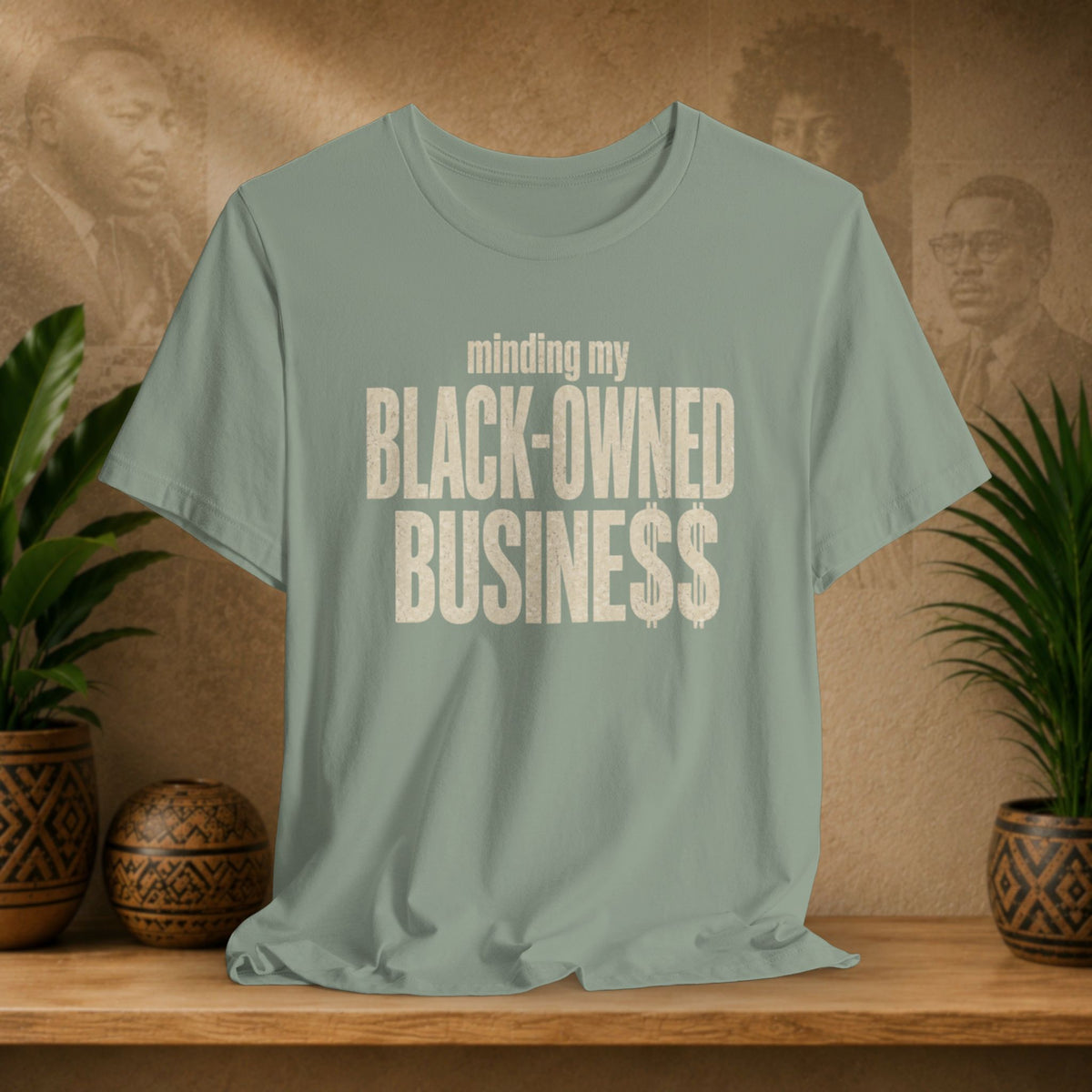A sage green graphic t-shirt displayed on a wooden shelf, flanked by potted plants. The shirt features the text 'minding my BLACK-OWNED BUSINESS$$' in distressed cream lettering.