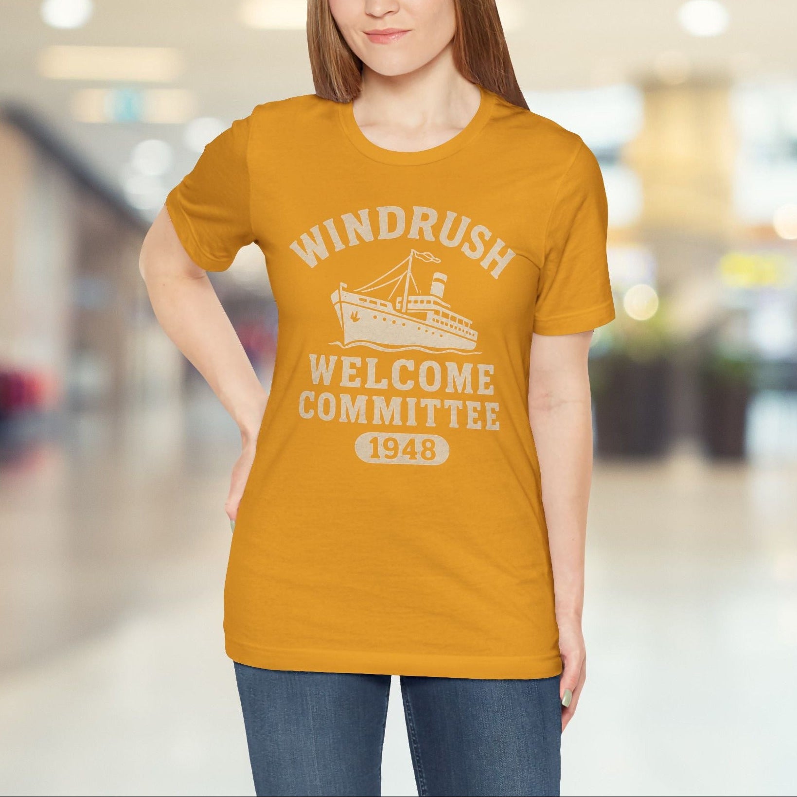 Woman wearing a mustard yellow t-shirt with 'Windrush Welcome Committee 1948' text and graphics in a blurred indoor setting.