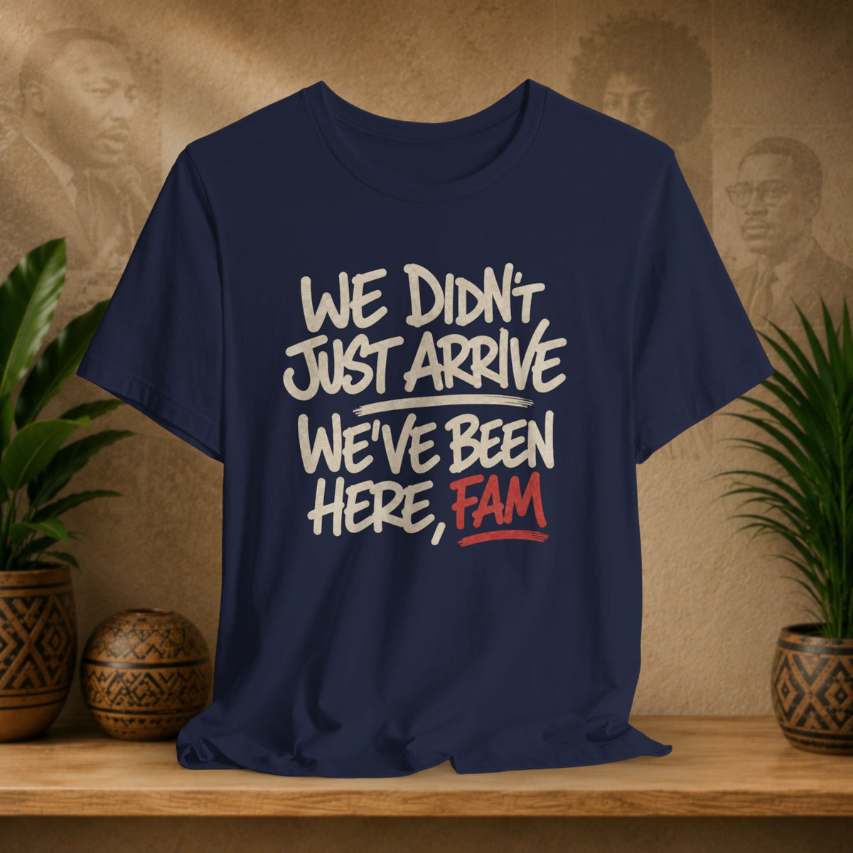 A navy blue graphic t-shirt displayed on a wooden shelf, flanked by potted plants. The shirt features a white and red printed slogan that reads 'WE DIDN'T JUST ARRIVE WE'VE BEEN HERE, FAM'.