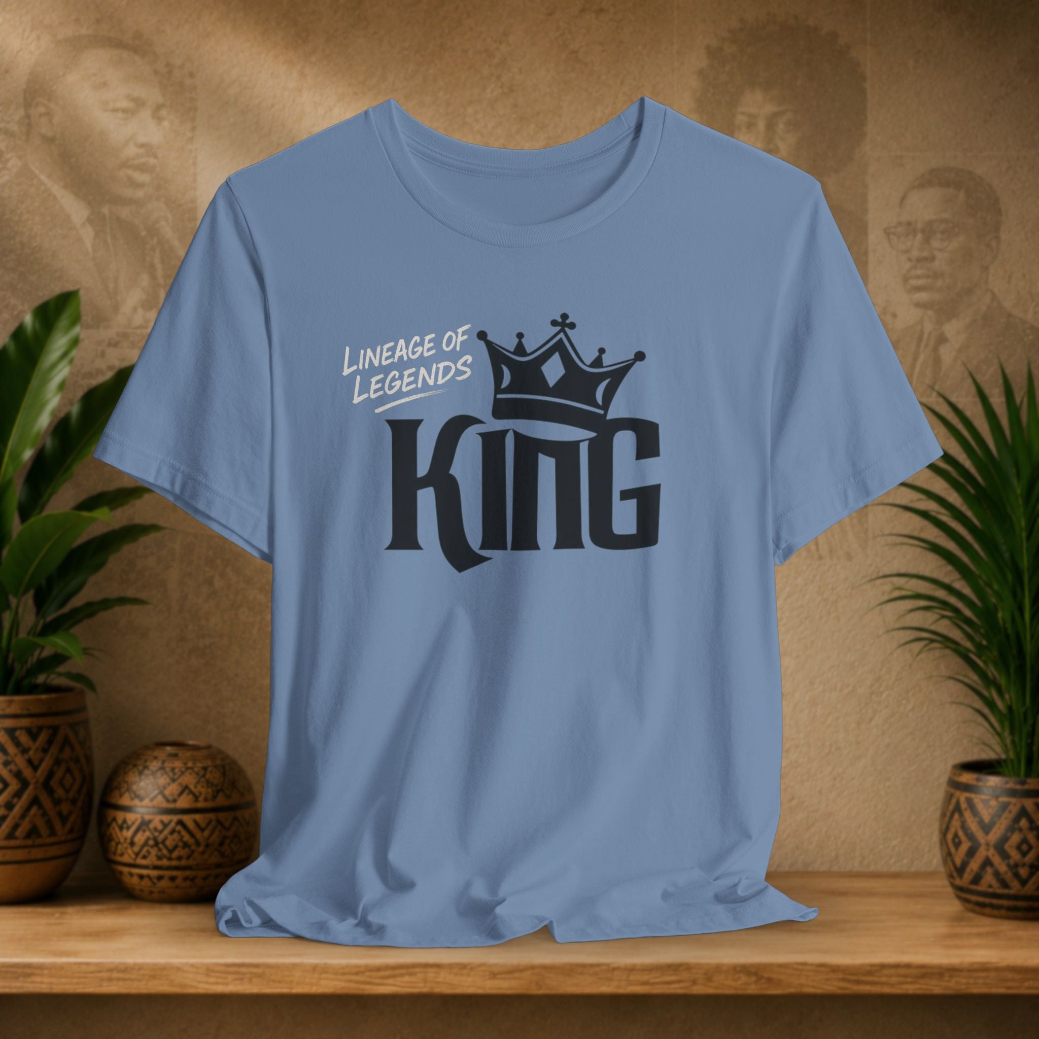 Light blue Lineage of Legends King graphic tee with black crown design, displayed on a shelf with plants and Afrocentric portraits