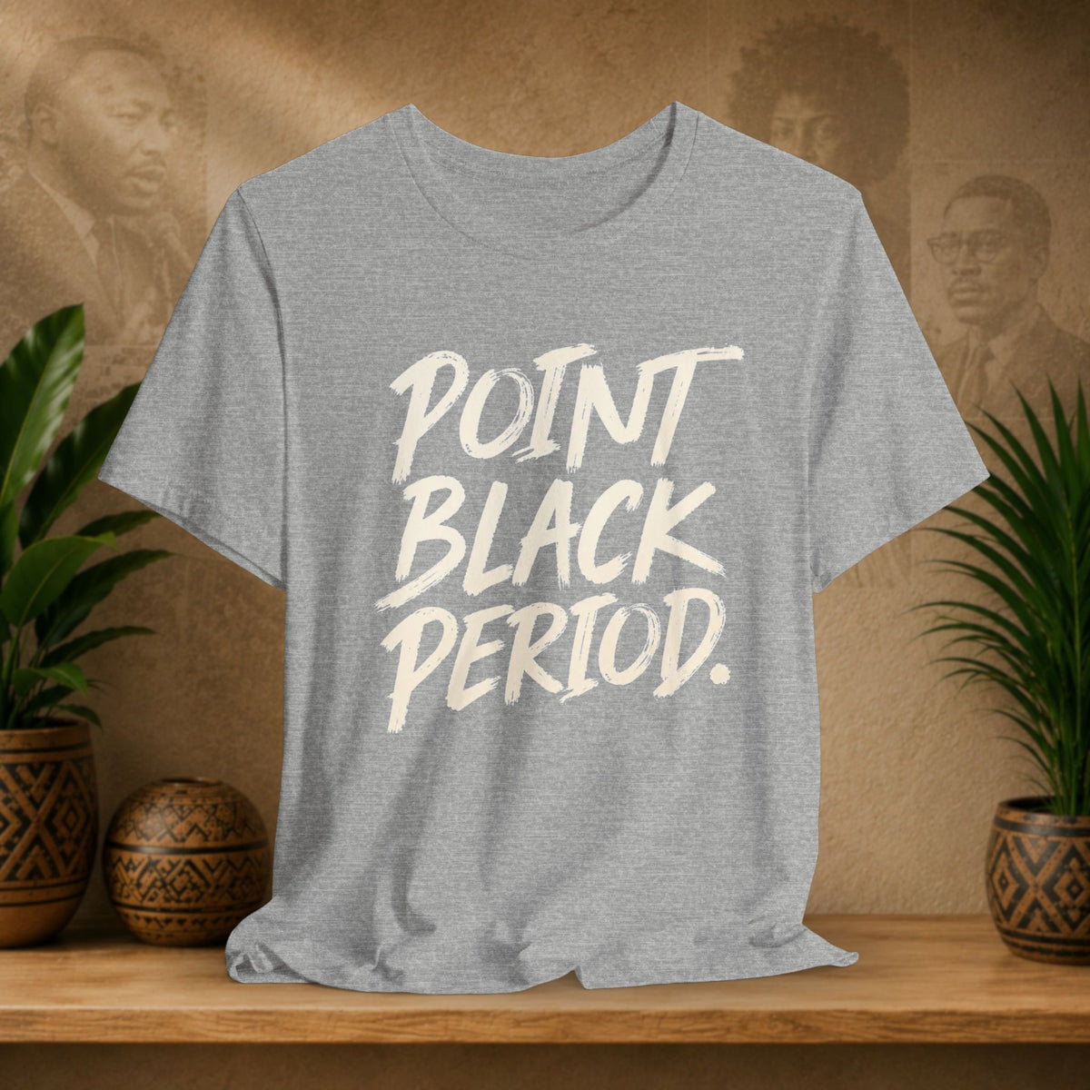 A heather gray short-sleeve t-shirt with the words 'POINT BLACK PERIOD.' printed in a large white brush-style font on the front, displayed on a wooden shelf with potted plants and a textured background.