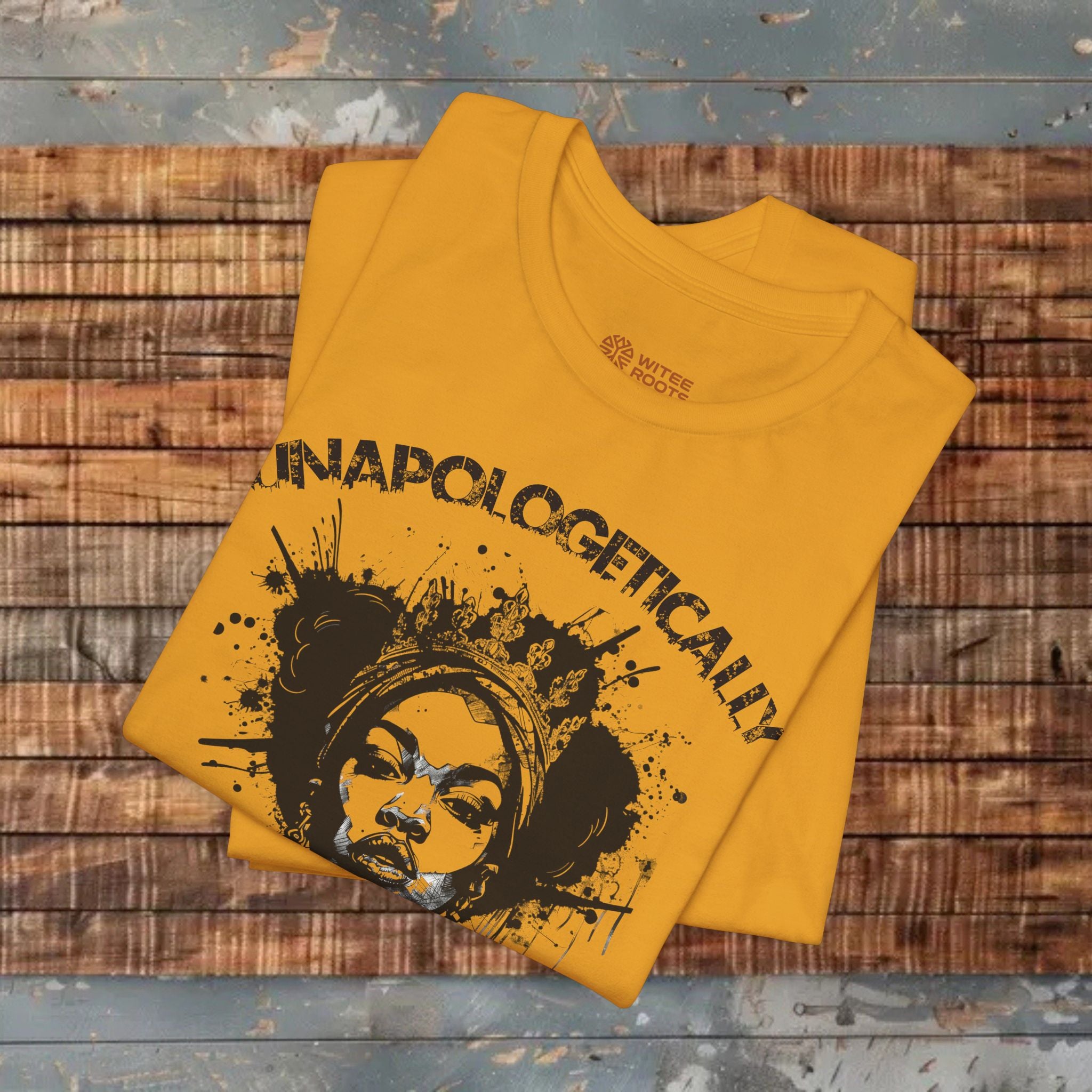 A folded mustard yellow t-shirt with a black graphic print reading 'UNAPOLOGETICALLY' and a stylized face, displayed on a rustic wooden plank surface.