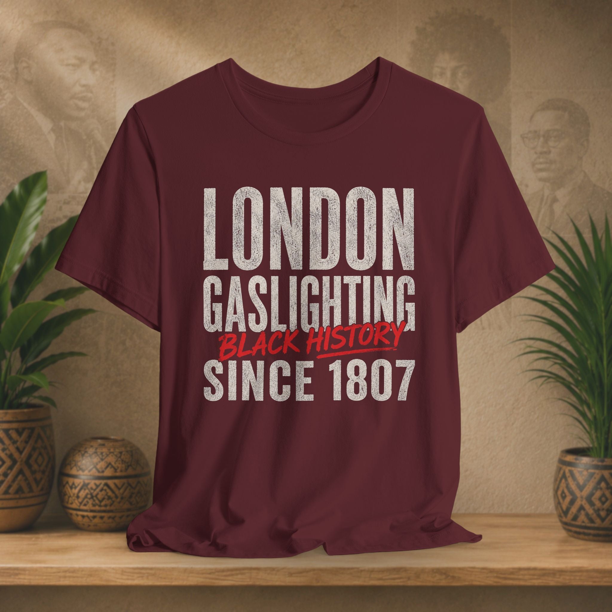 Maroon t-shirt with 'London Gaslighting Black History Since 1807' text on a wooden surface with plants and a blurred background.