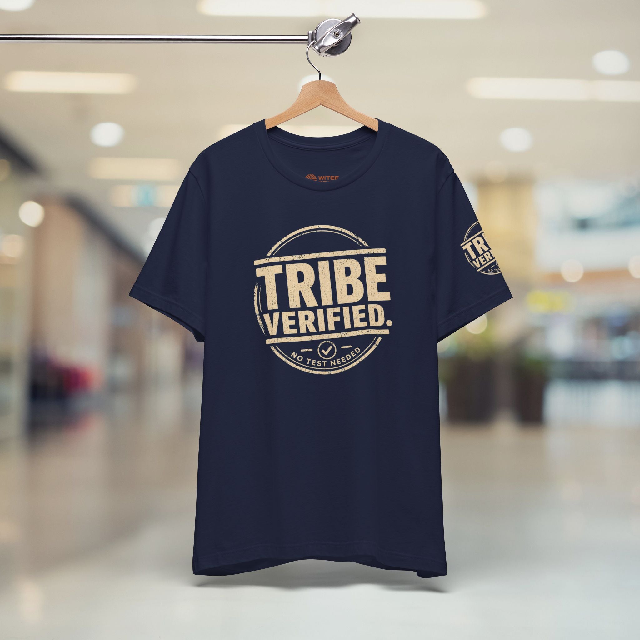 Navy blue t-shirt with 'TRIBE VERIFIED' text on a hanger in a blurred indoor setting
