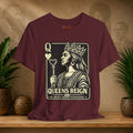Maroon t-shirt with a graphic design of a queen and text on a wooden surface with plants and a textured wall in the background.