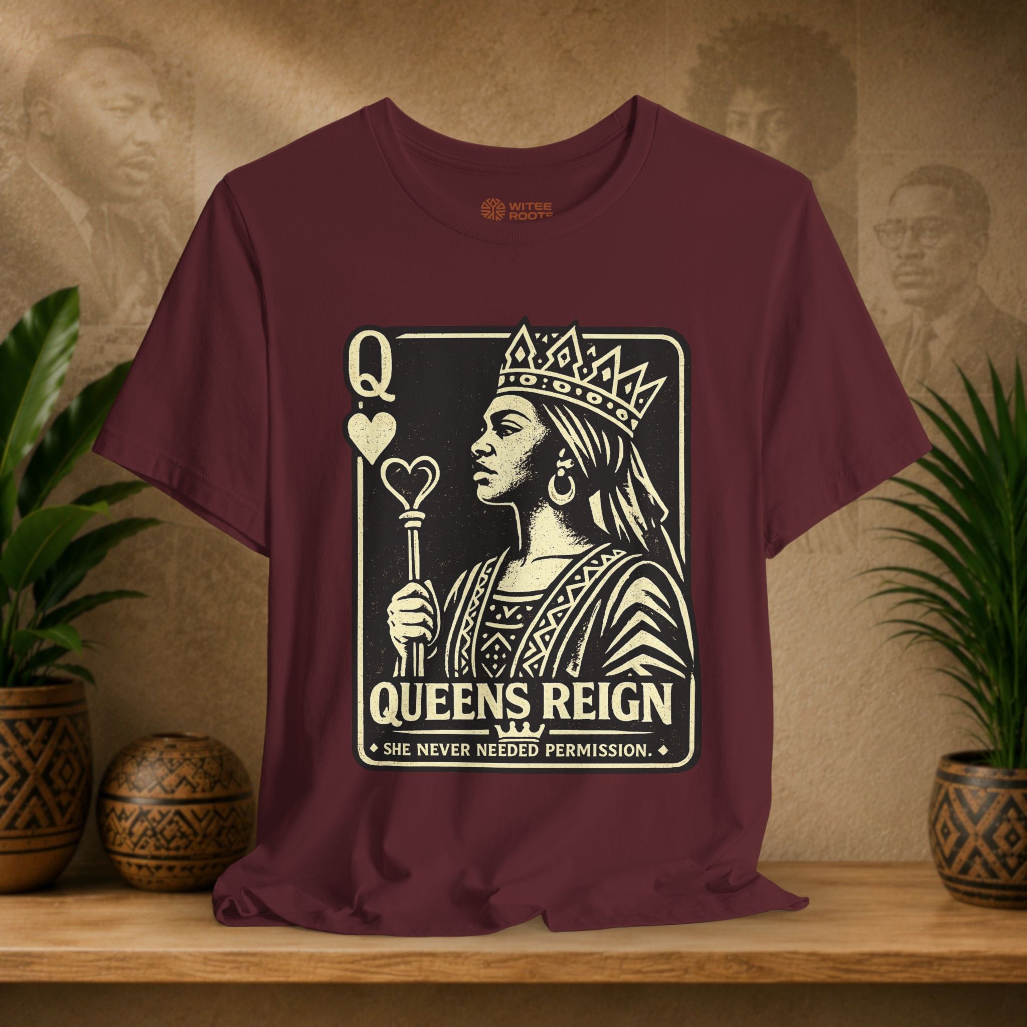Maroon t-shirt with a graphic design of a queen and text on a wooden surface with plants and a textured wall in the background.